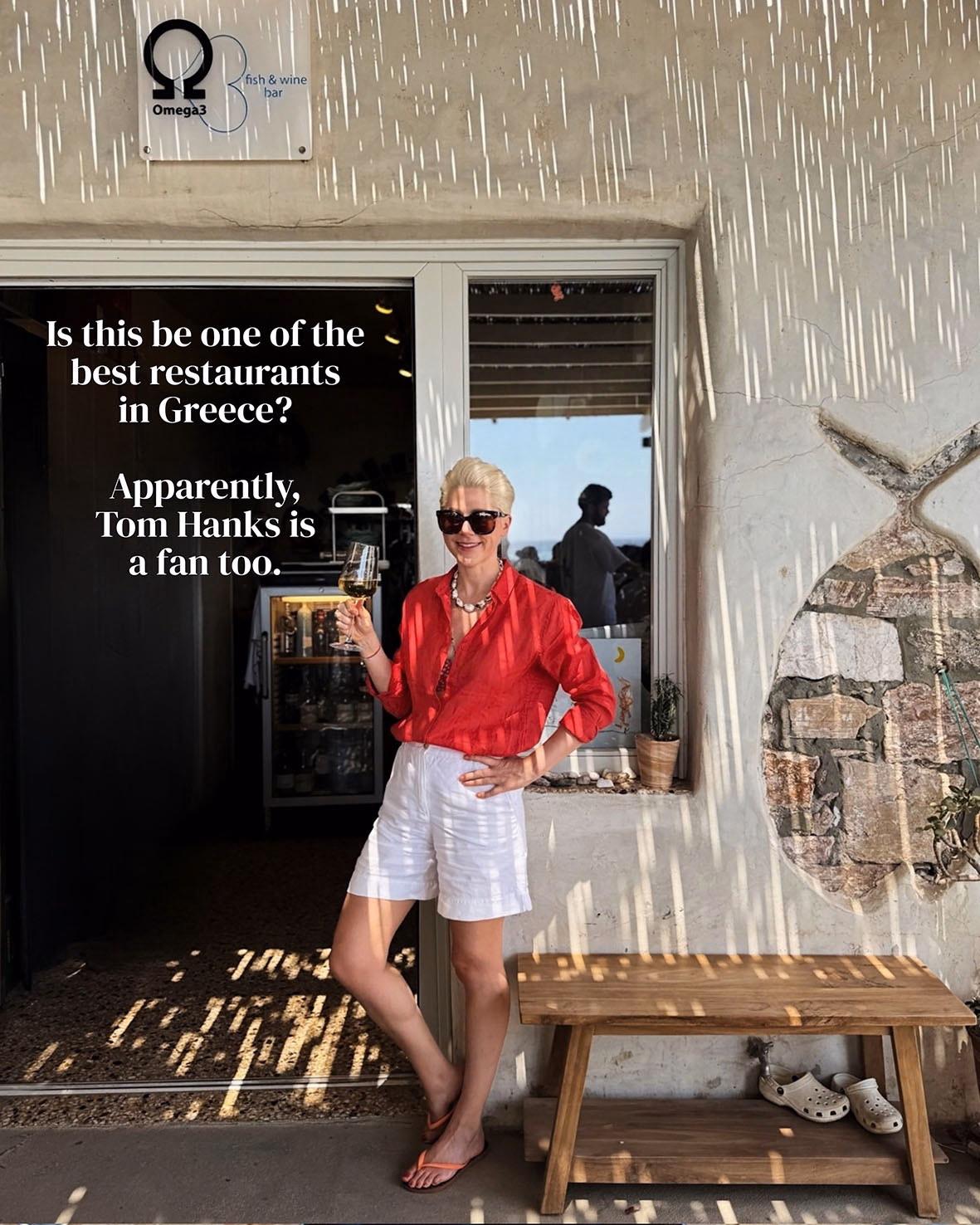 Woman in red linen shirt and white shorts holds a glass of white wine outside Omega3 fish and wine bar, Sifnos.