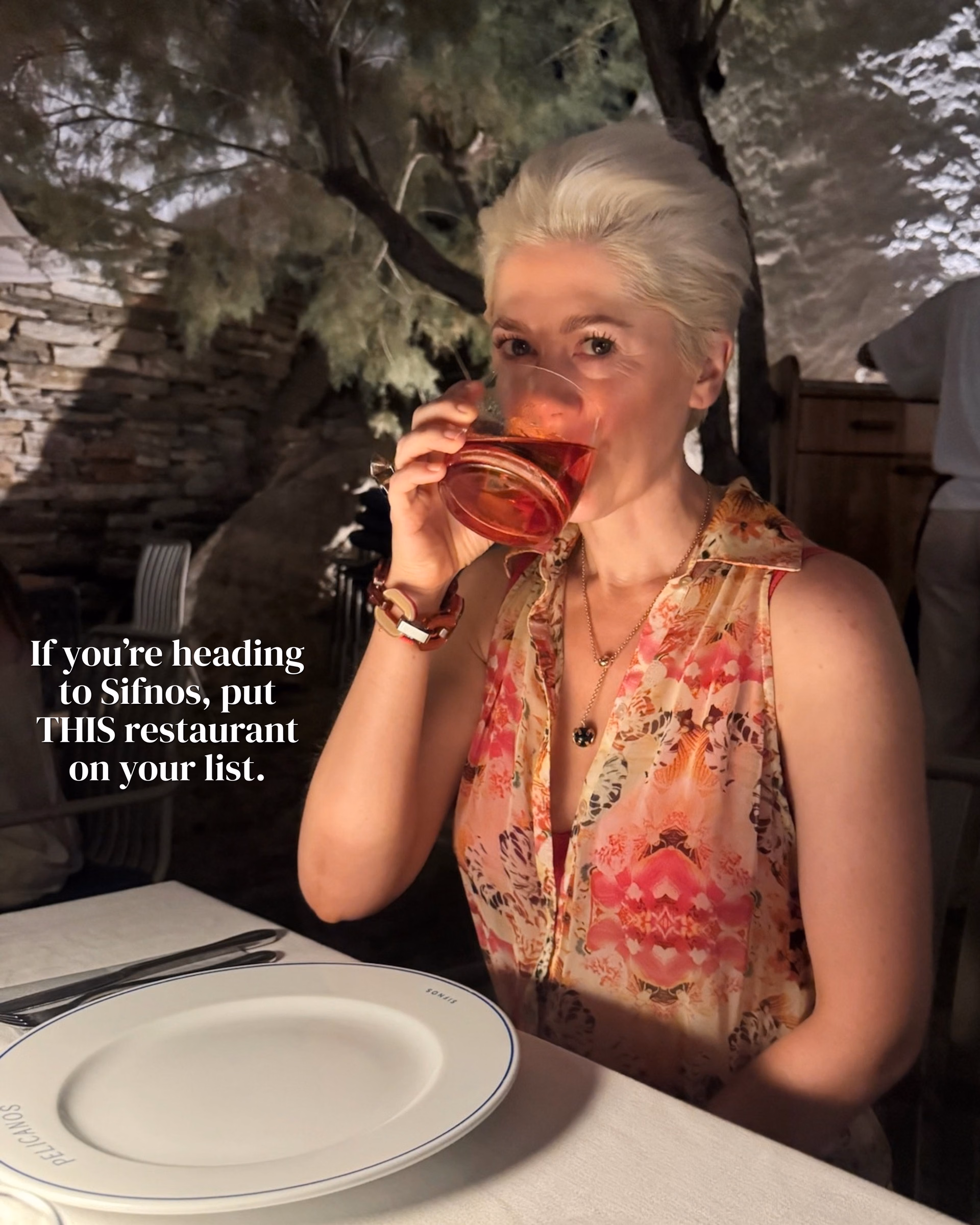 Woman with short platinum hair in a sleeveless floral top sipping red wine at a table with a Pelicanos plate, under a tree.