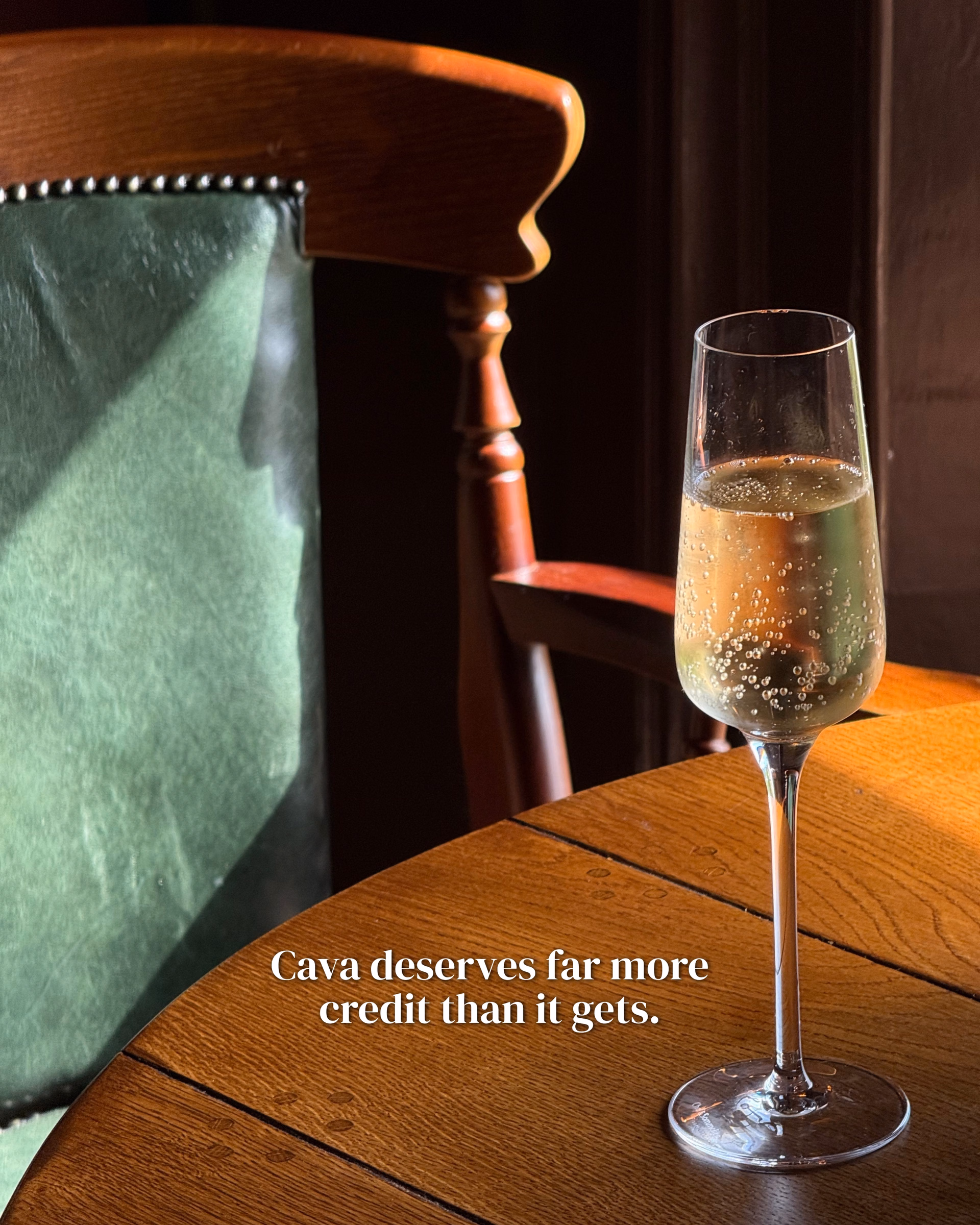 A flute of sparkling wine on a wooden table beside a green velvet chair with studded trim, lit by warm sunlight.