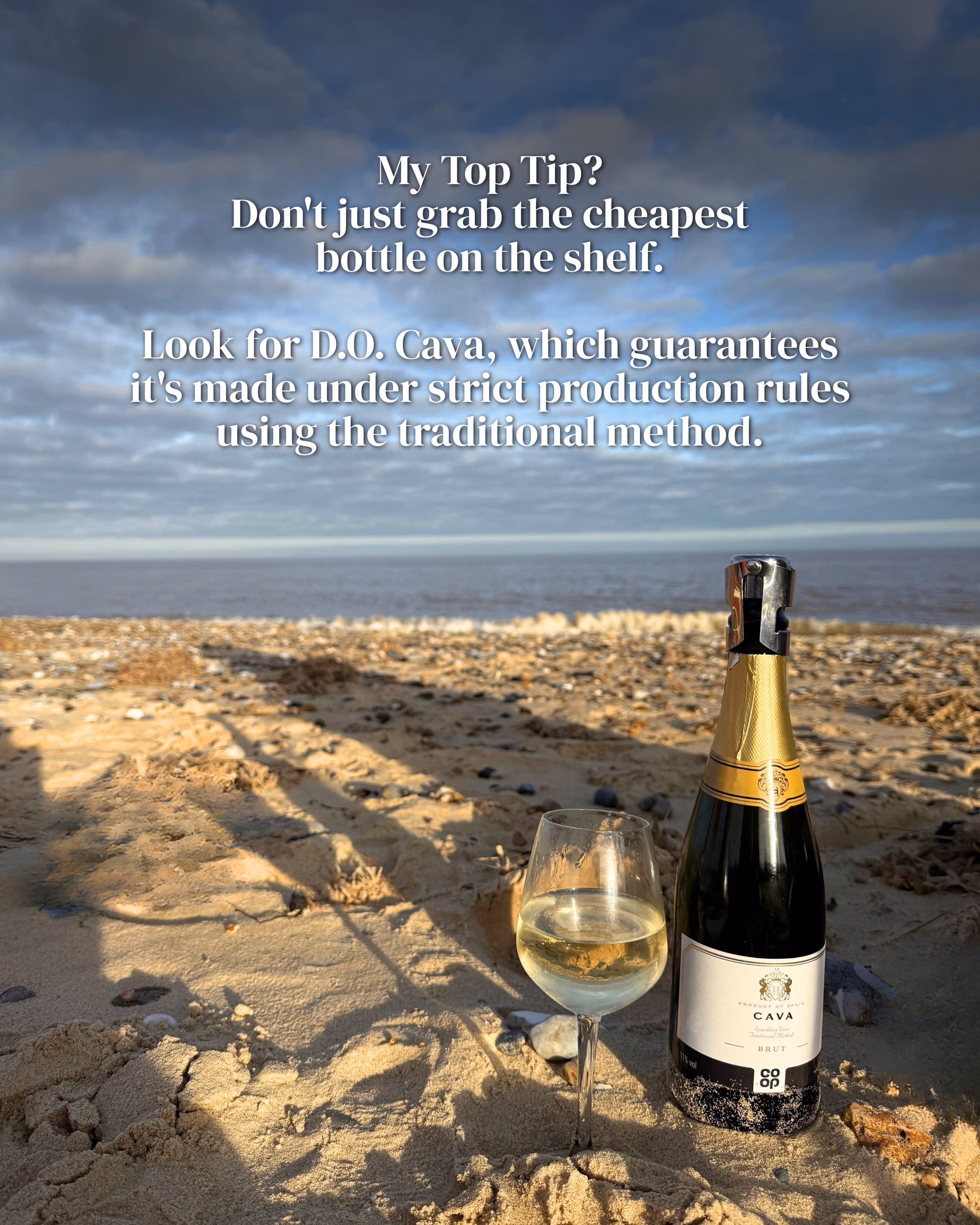 Bottle of Co-op Cava Brut beside a glass of sparkling wine on a sandy beach at sunset, with text overlay.