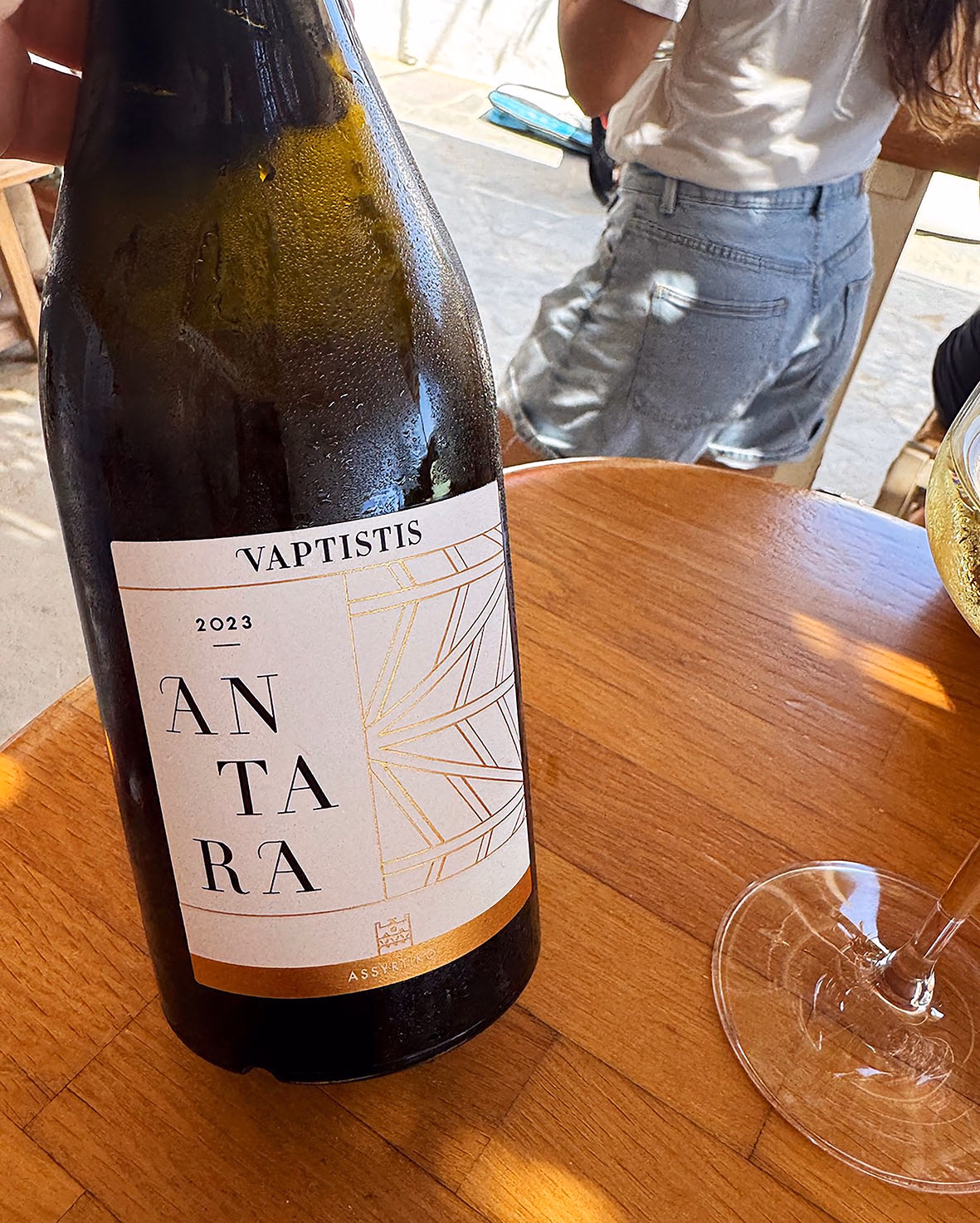 Bottle of Vaptistis Antara 2023 wine, beaded with condensation, on a wooden table beside a glass, with diners in the background
