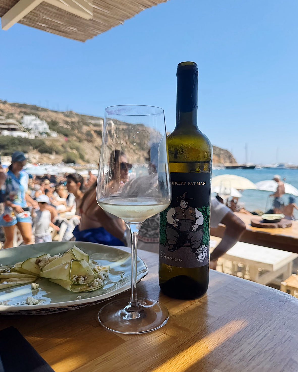 Glass of white wine beside a bottle of Sheriff Fatman Assyrtiko and a plate of zucchini rolls with crumbled cheese, overlooking a beach.