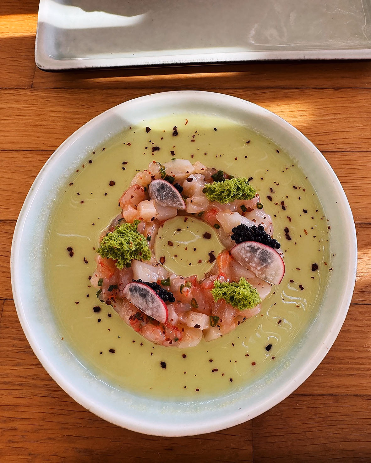 Raw fish tartare arranged in a ring on a pale green chilled soup, topped with radish slices, caviar and green herb crumbs.