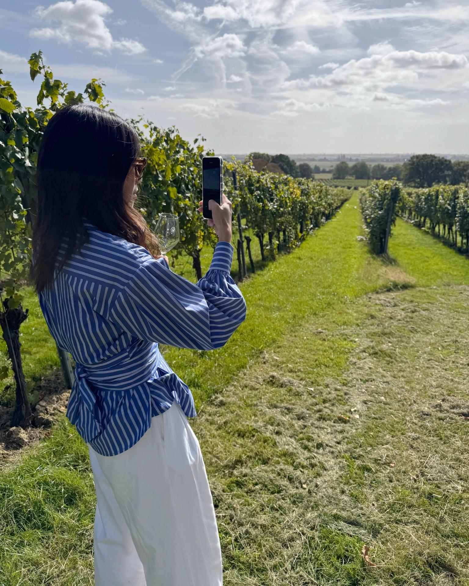Woman in a blue striped shirt and white trousers photographs rows of green vines with her phone, holding a wine glass.