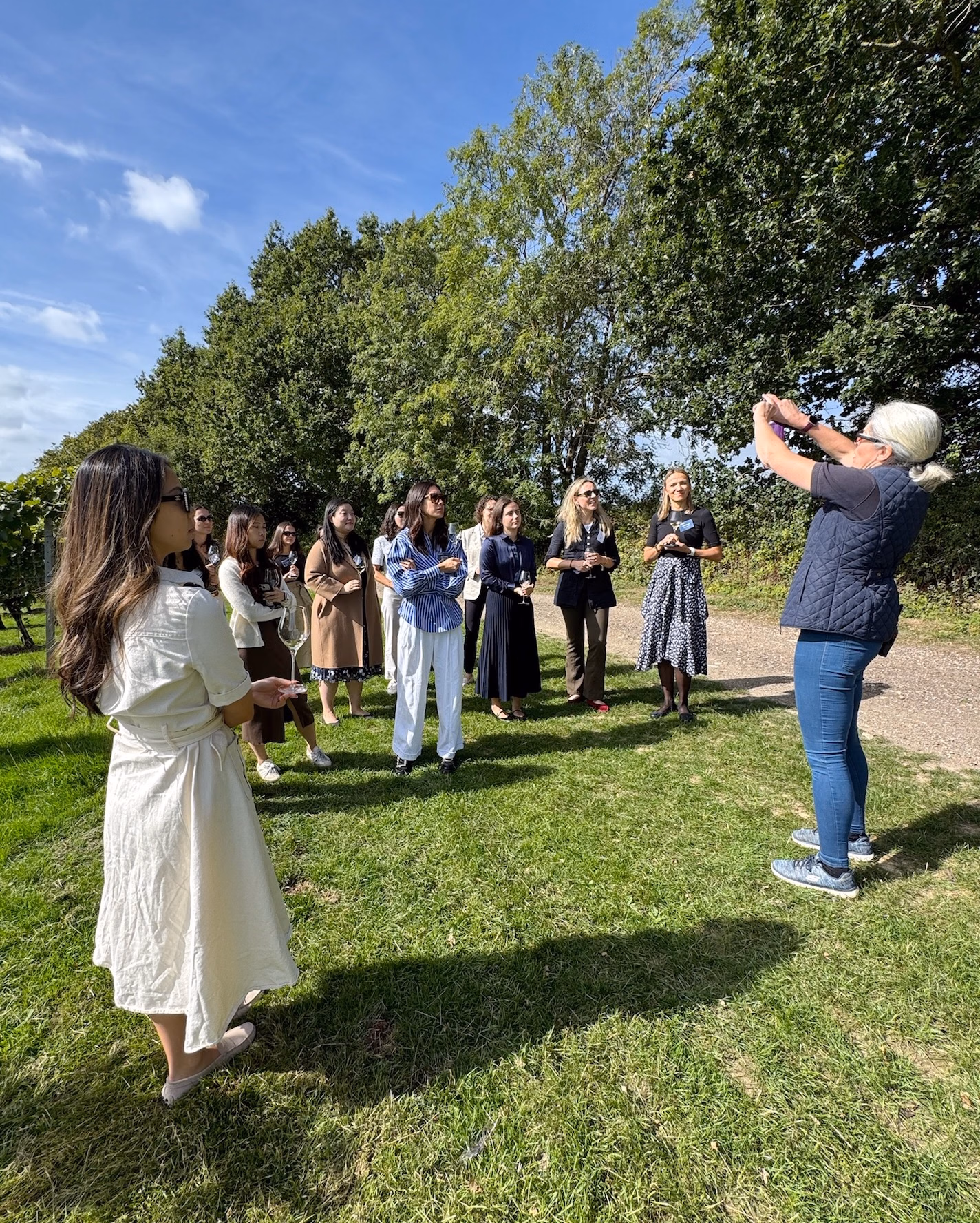 Group of women on a grassy vineyard path in summer sunshine, one with white hair photographing the group on her phone.