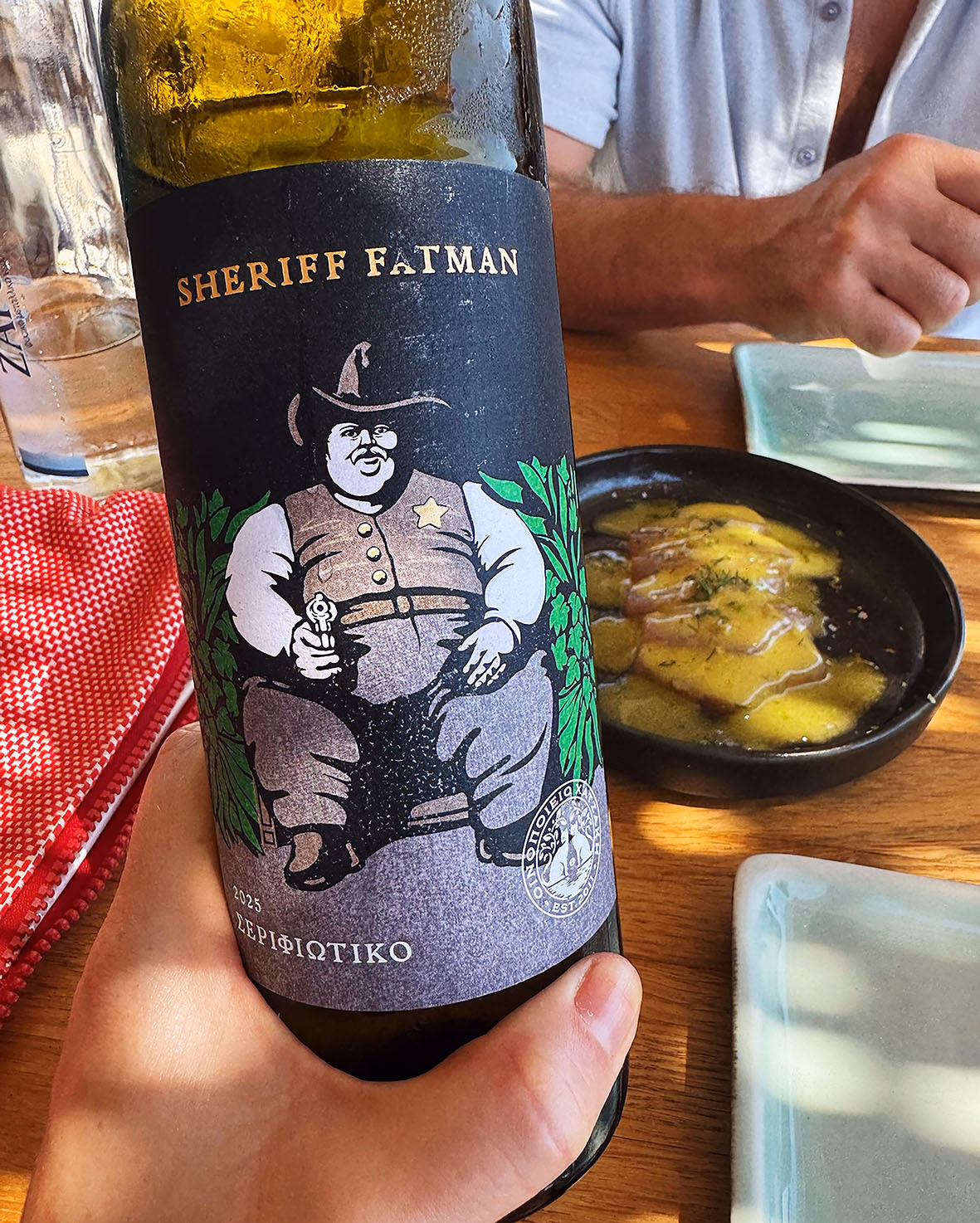 Hand holding a bottle of Sheriff Fatman 2025 wine with a cartoon sheriff label, beside a dish of fish collar at a wooden table.