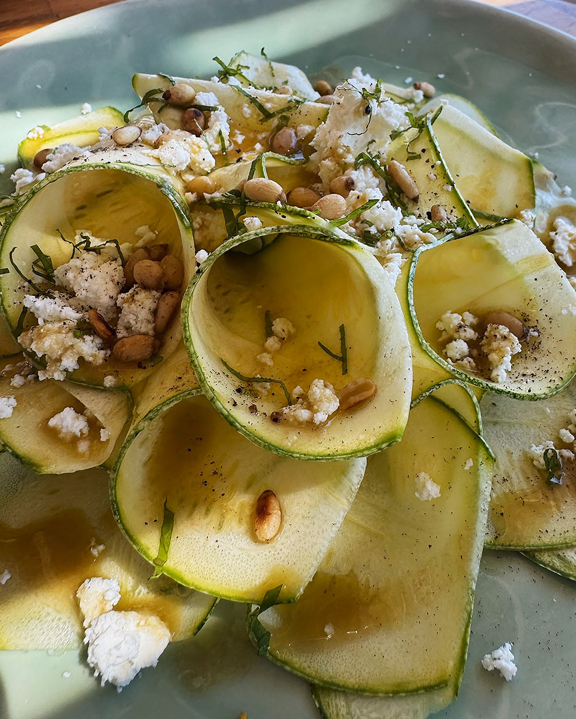 Shaved courgette ribbons curled on a pale plate, dressed with olive oil, crumbled cheese, pine nuts and fresh herbs.