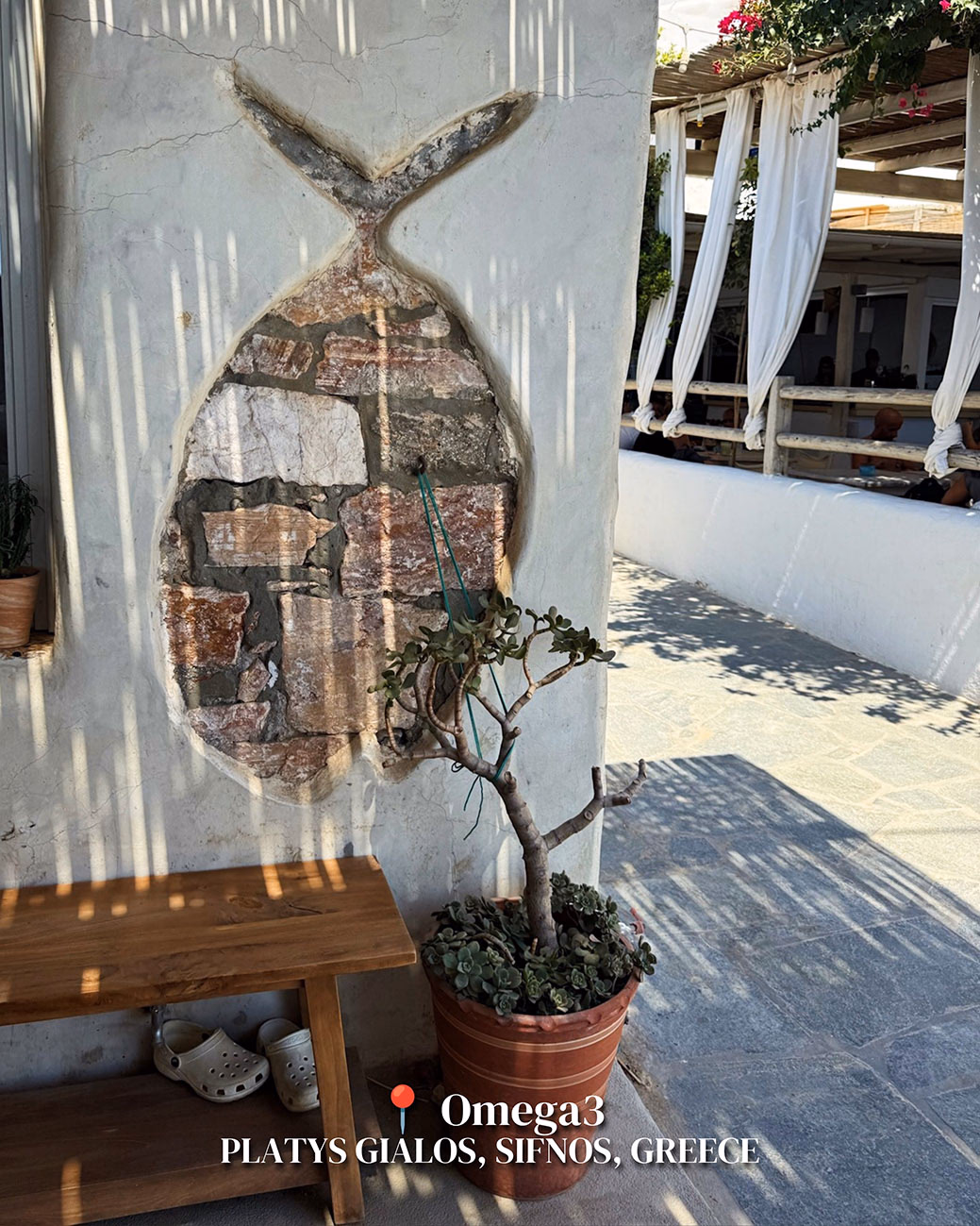 Whitewashed wall with a fish-shaped stone mosaic, a potted jade plant, a wooden bench and sandals at Omega3 on Sifnos