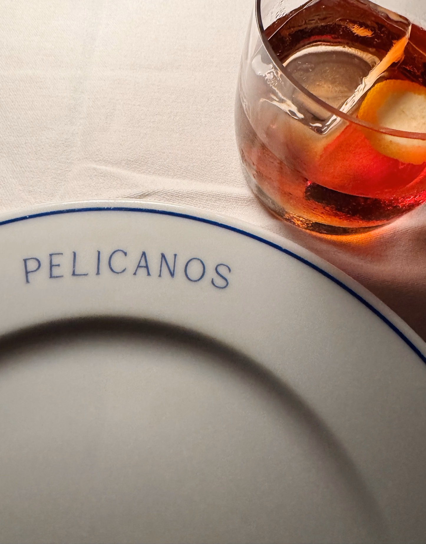 White plate with blue rim reading "PELICANOS" beside a glass of red cocktail garnished with an orange peel on white linen.