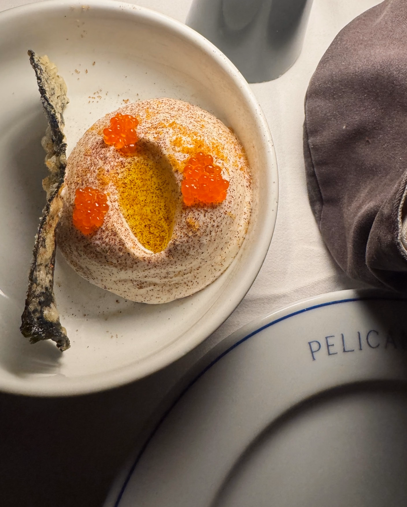Dish of whipped creamy mousse dusted with paprika, topped with orange trout roe and golden oil, beside a crisp fried fish skin.