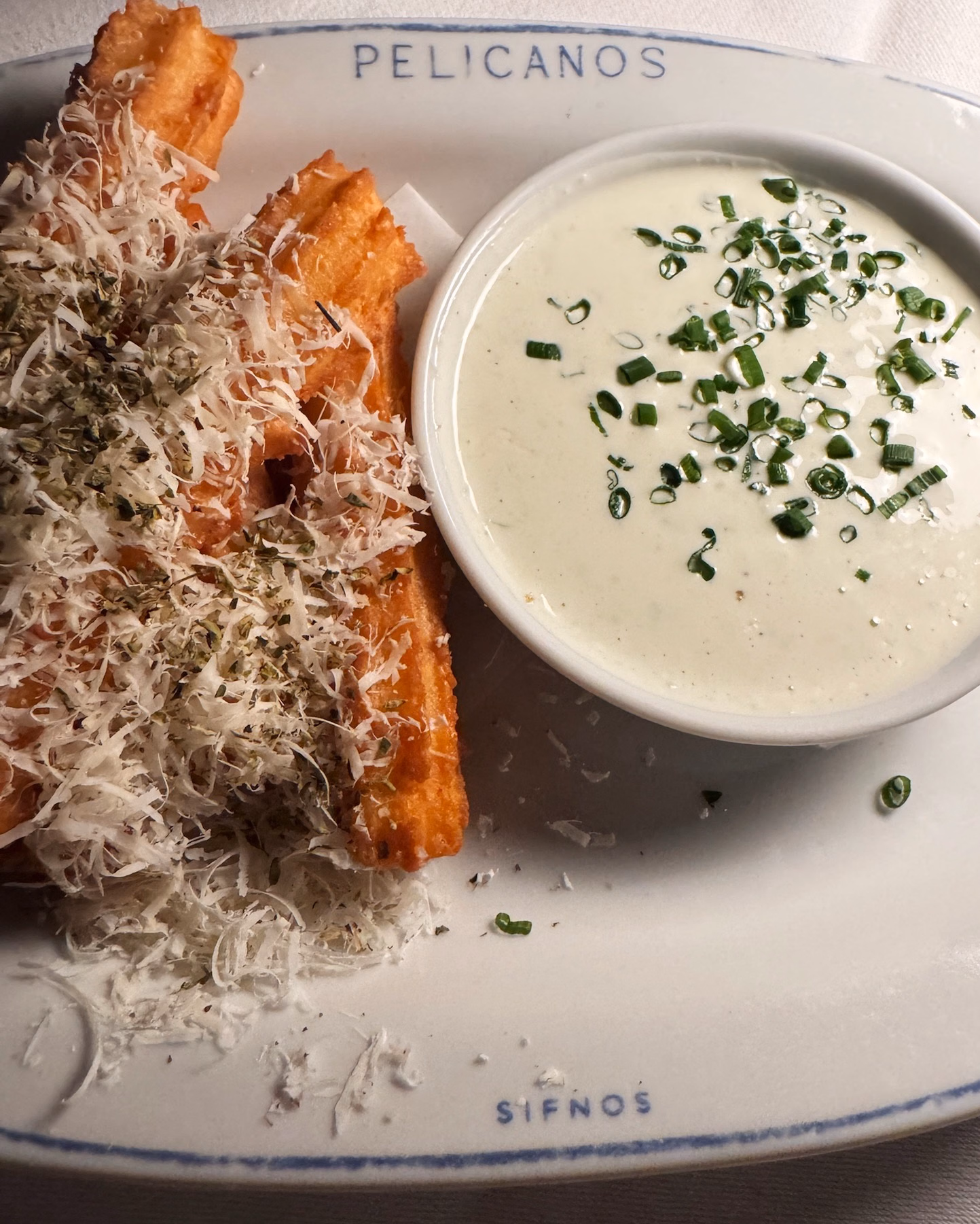 Crispy fried potato wedges topped with grated cheese and herbs, served with a creamy white dip garnished with chives, on a Pelicanos plate.