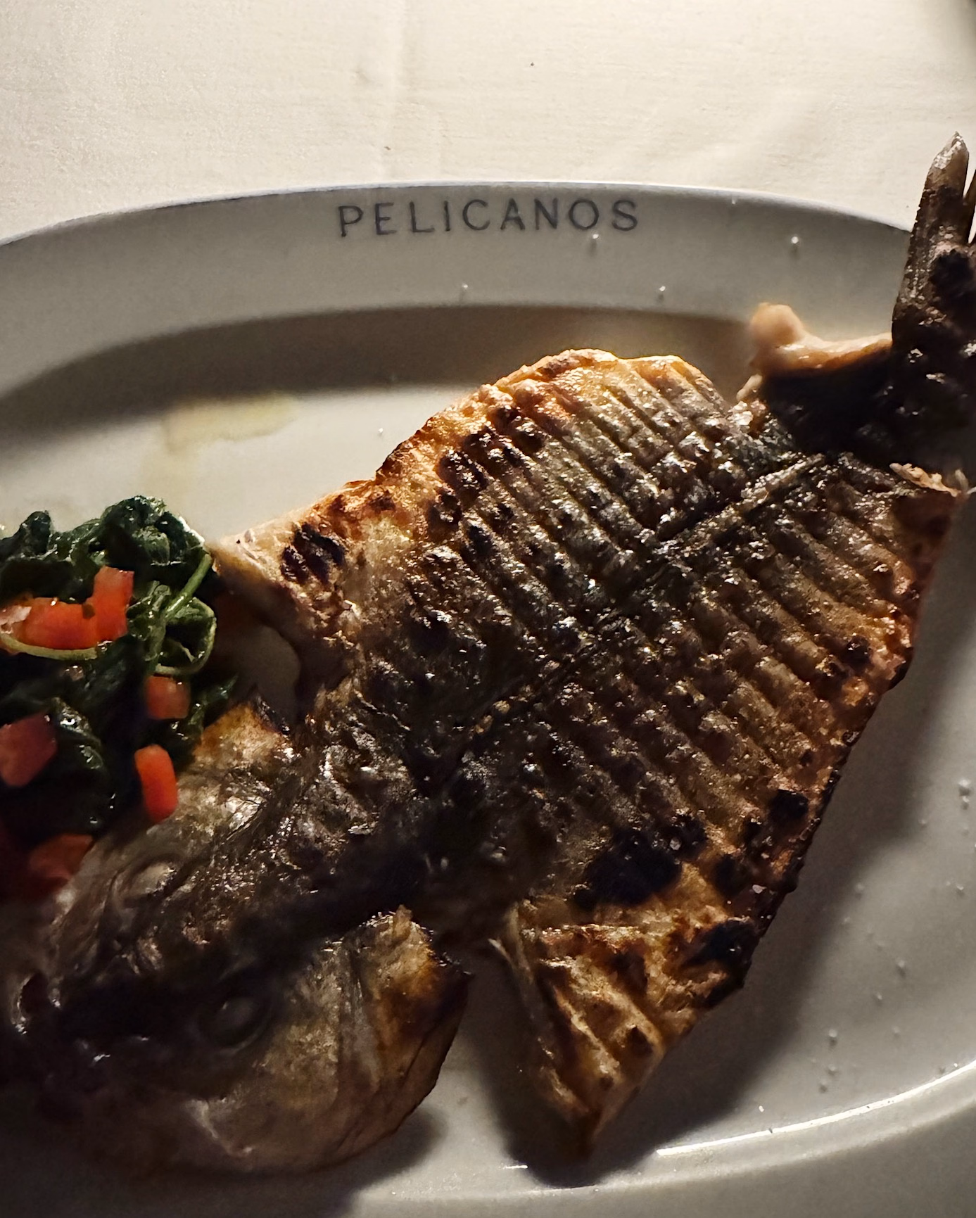 Grilled whole sea bream with charred grill marks on a white Pelicanos plate, served with sautéed spinach and diced tomato.
