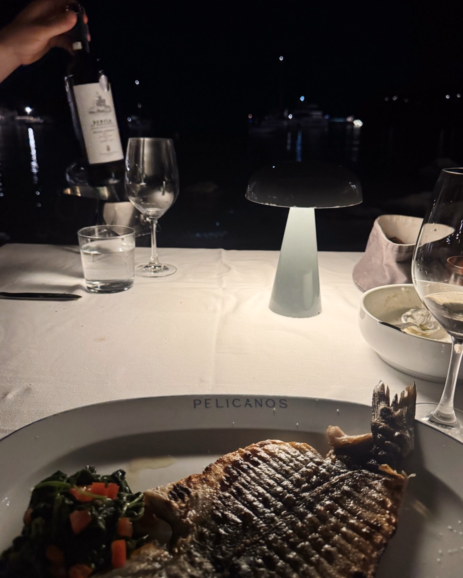 Grilled fish with spinach and tomato on a white plate at a night-time table, with wine, a candle-style lamp and the sea behind.