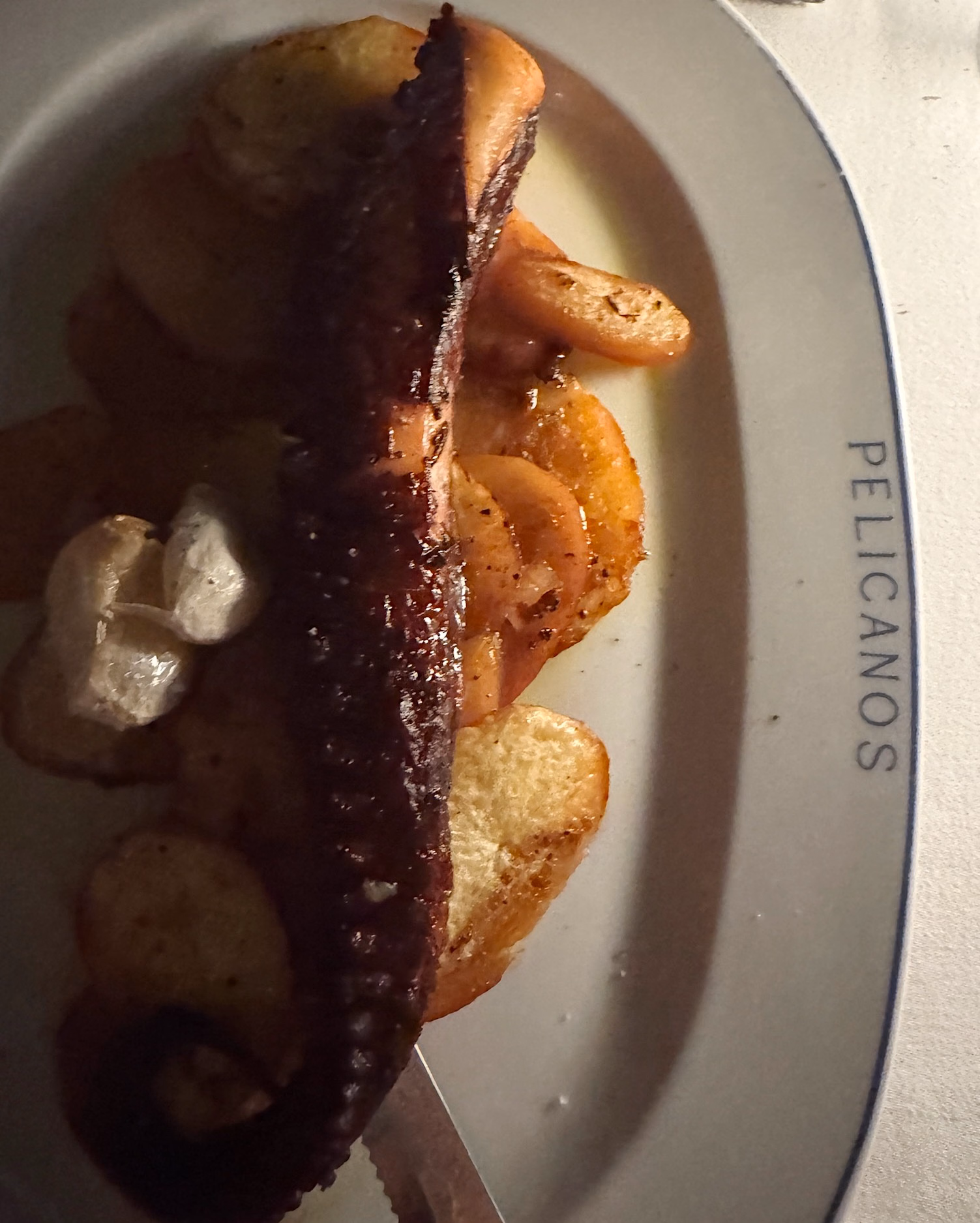Grilled octopus tentacle with crispy potato slices on a white plate marked "Pelicanos", with a knife beside it.