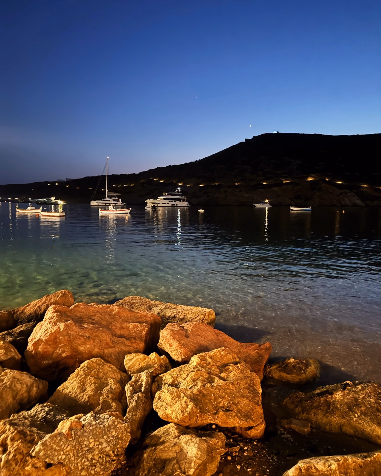 Dusk over a calm Aegean bay, with glowing rocks in the foreground, moored yachts and fishing boats, and a lit hillside.