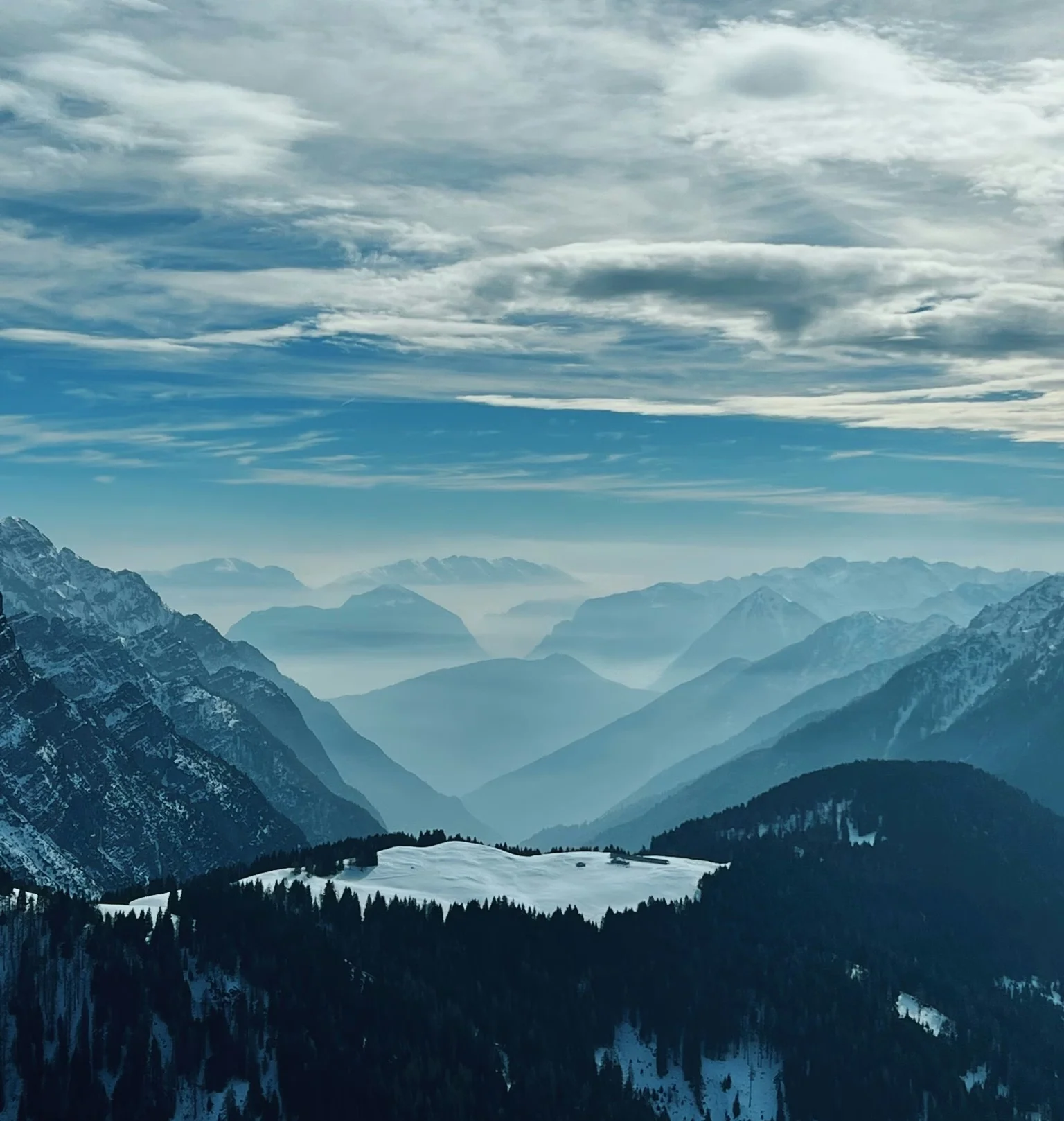 Snow-covered Alpine mountains and forest beneath a dramatic cloud-filled sky