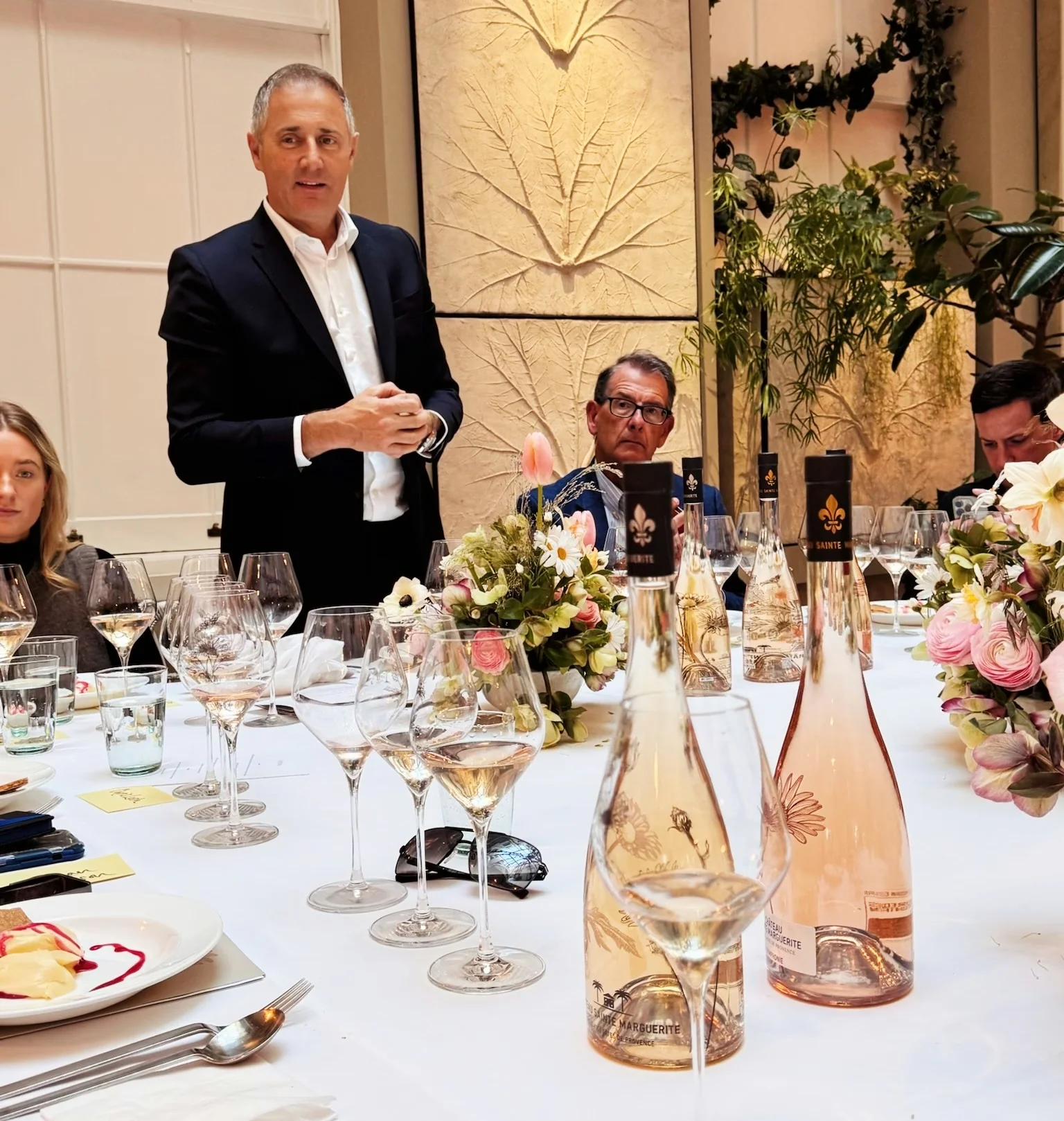 A man speaks at a dining table set with rosé wine, glasses, flowers and plates of food.