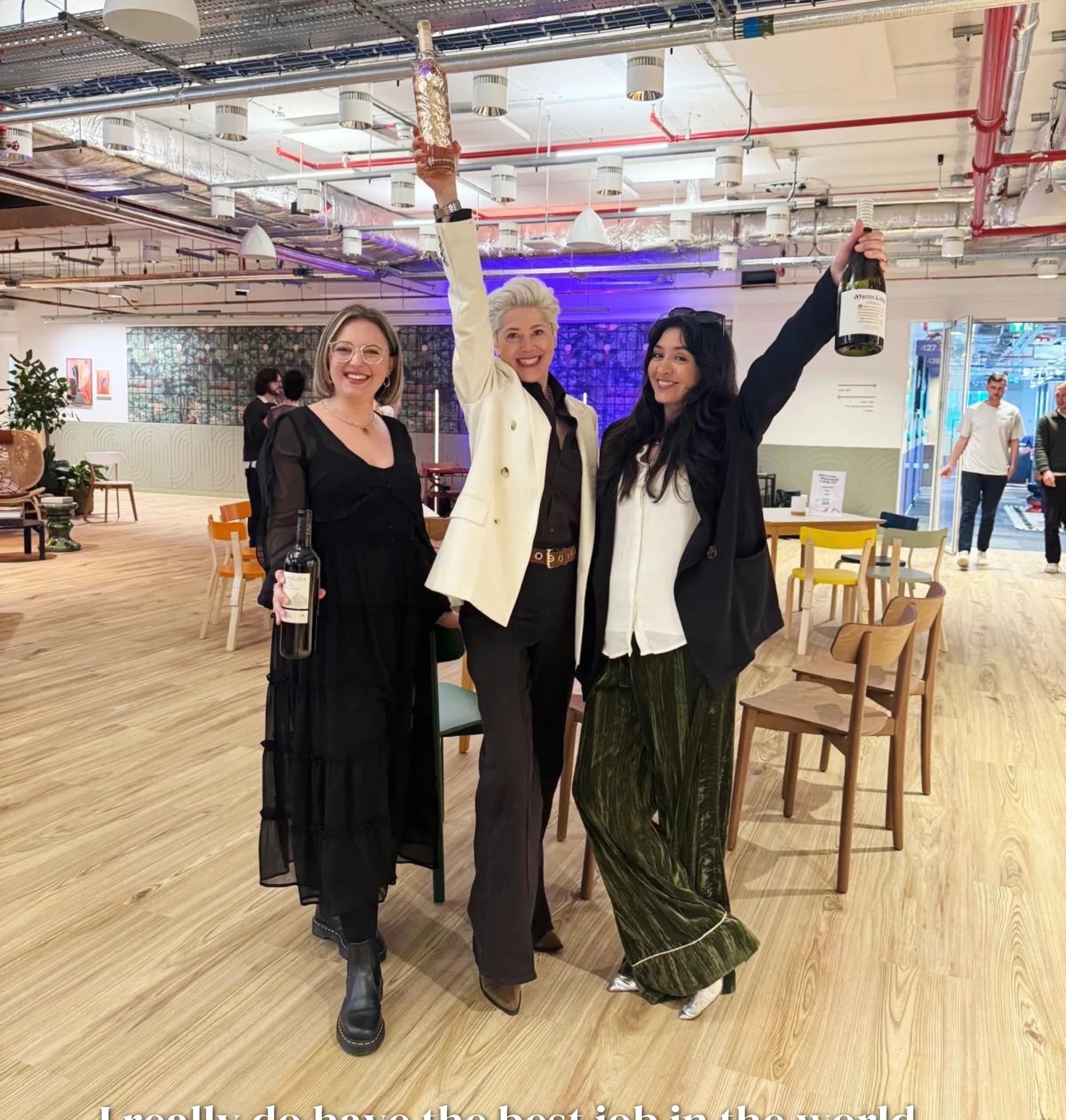 Three women smiling and raising wine bottles in a bright event space