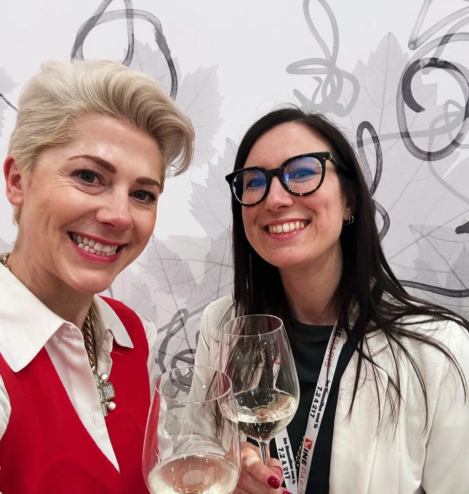 Two women smiling at a wine event while holding glasses of white wine