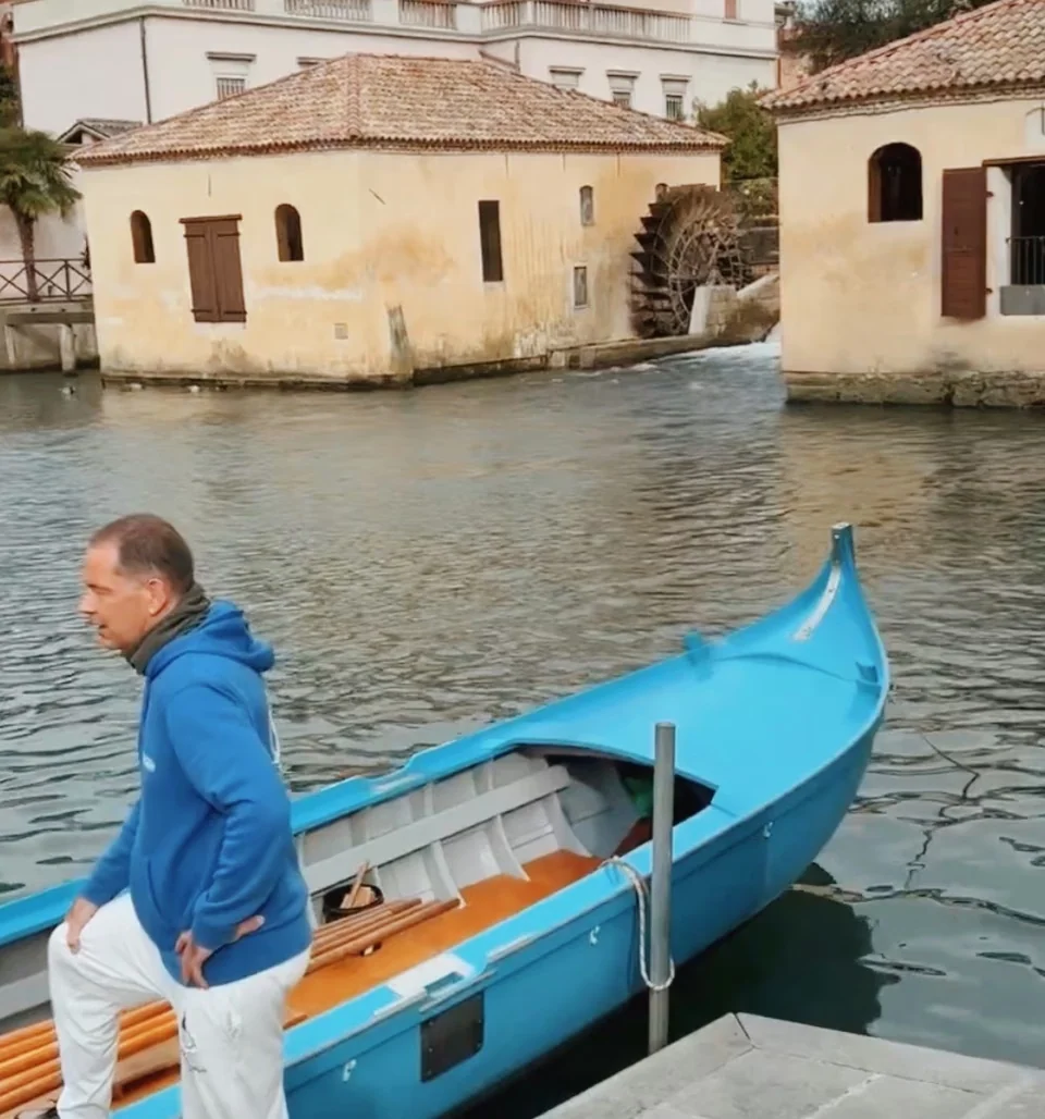 Man beside a bright blue boat on a canal, with historic cream buildings and a waterwheel behind