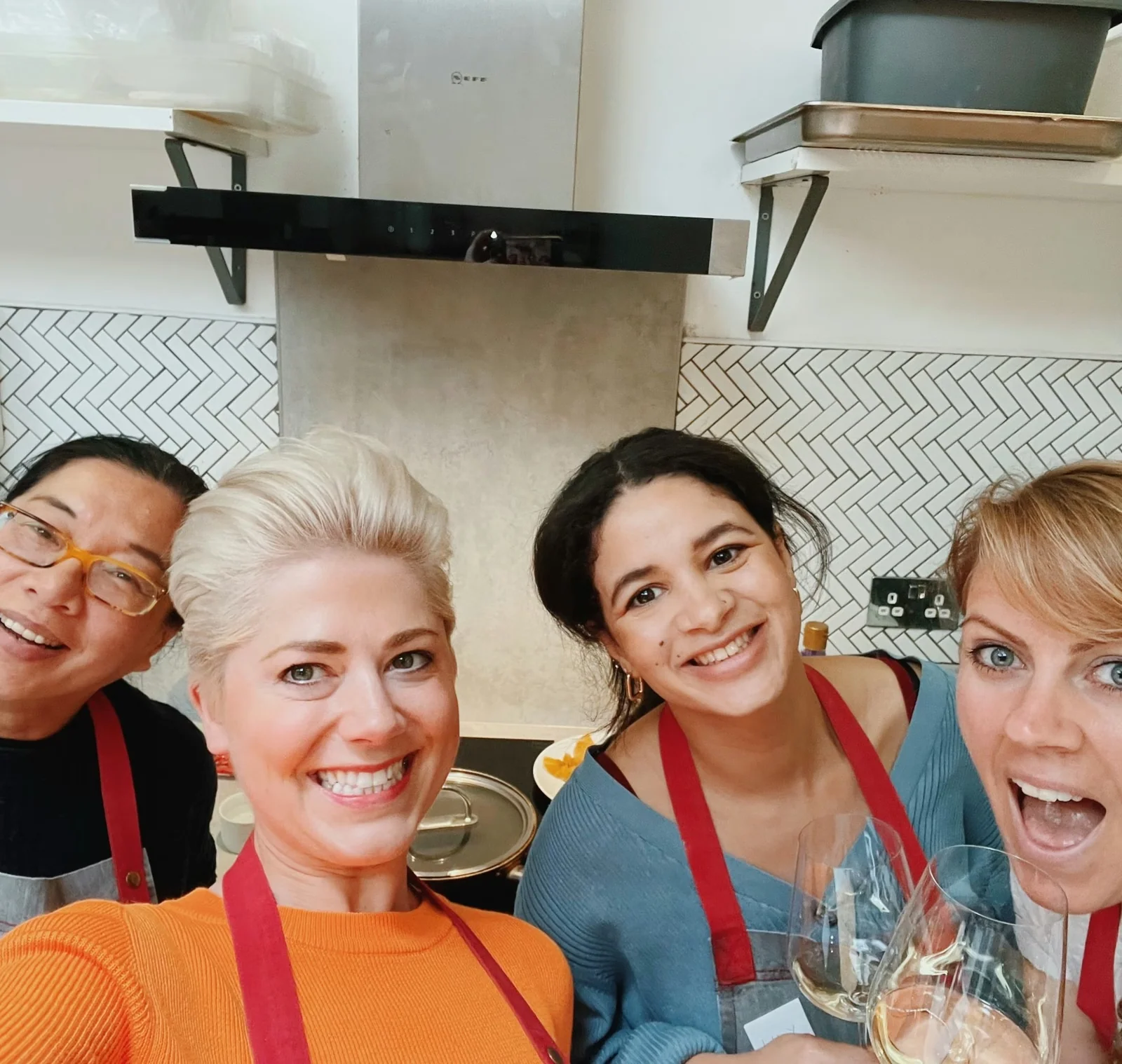 Four women in red aprons smile for a selfie in a tiled kitchen, with glasses of white wine