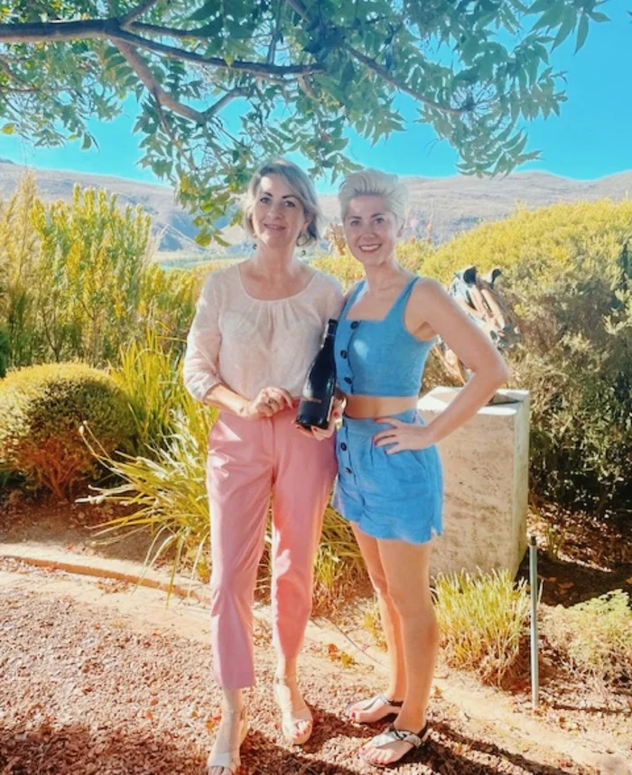 Two women stand beneath a tree in a sunny landscaped vineyard, one holding a bottle of wine.
