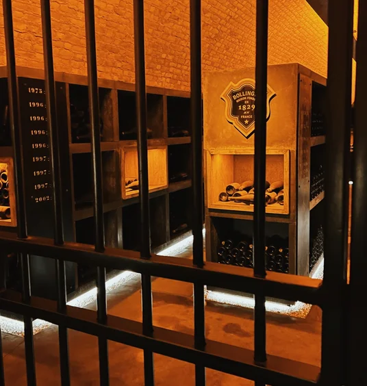 Bollinger Champagne bottles stored in a warmly lit underground cellar behind black iron bars