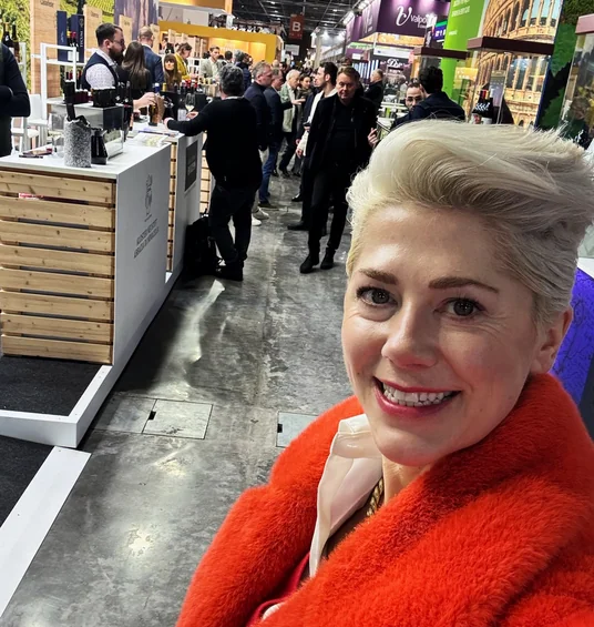 Elizabeth Hawthornthwaite at a busy wine exhibition, wearing a bright red coat beside tasting stands