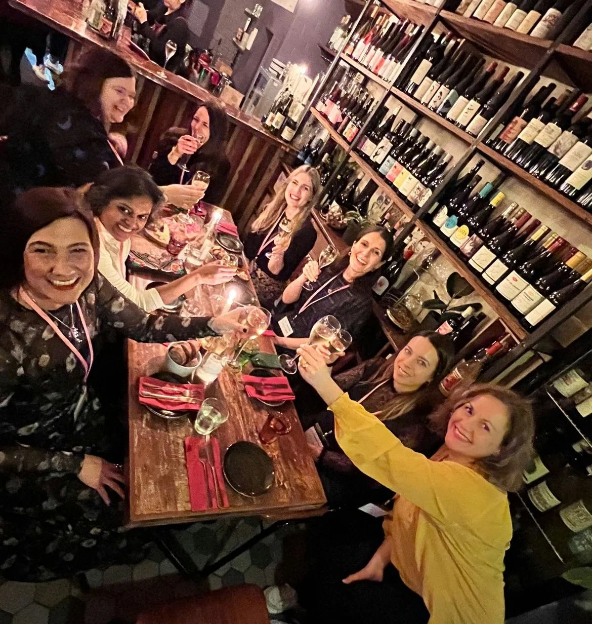 Women raising wine glasses around a table at Lady of the Grapes, with wine bottles lining the shelves
