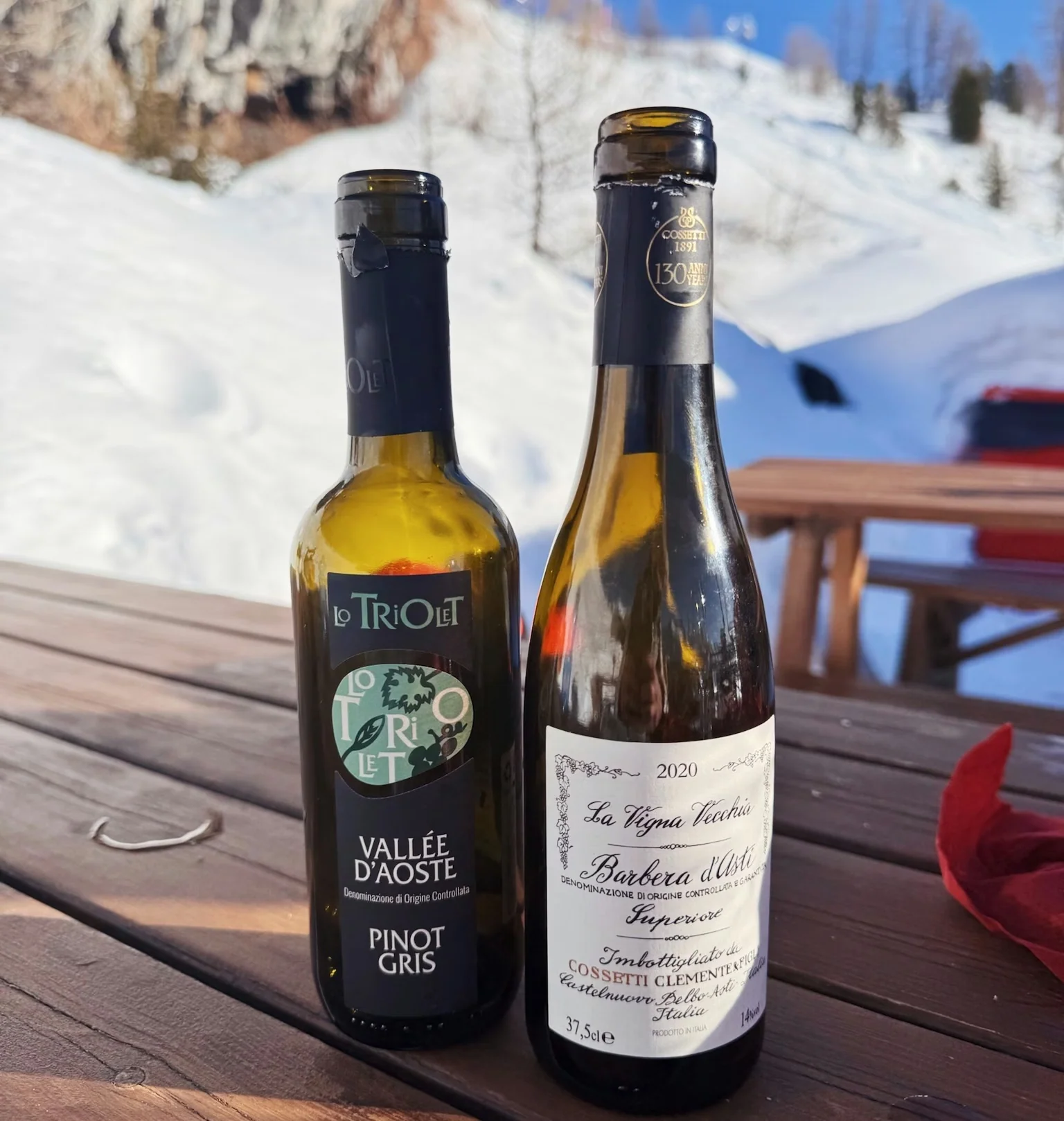 Two bottles of Italian wine on a wooden table overlooking a snowy ski slope in Champoluc