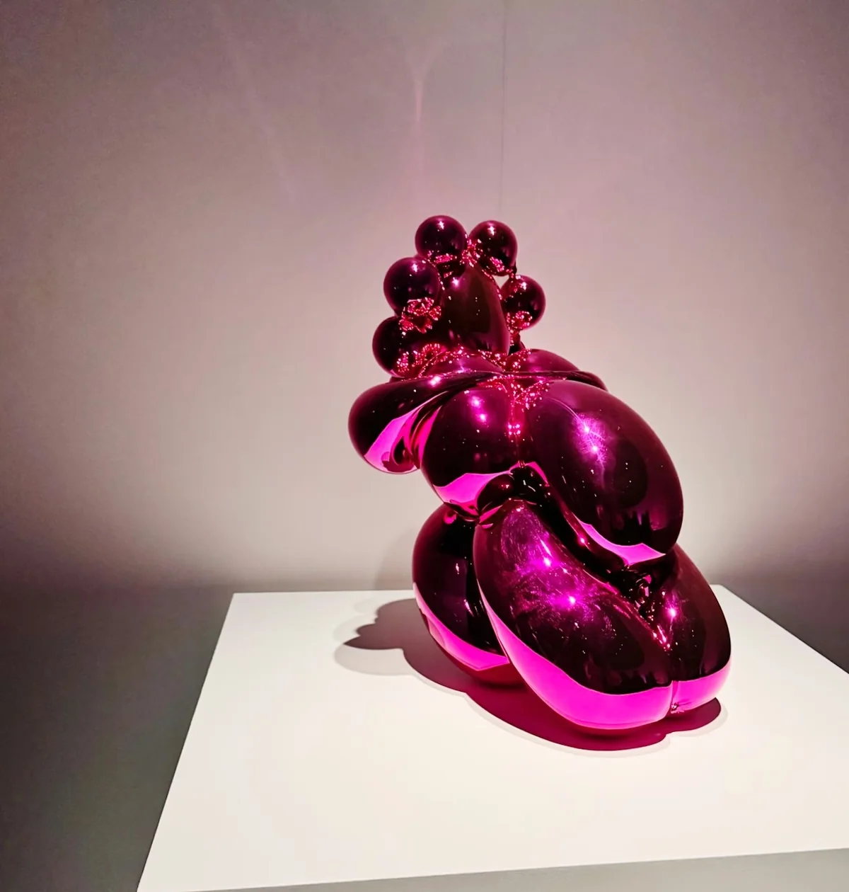 Glossy magenta abstract sculpture displayed on a white plinth in a dim gallery