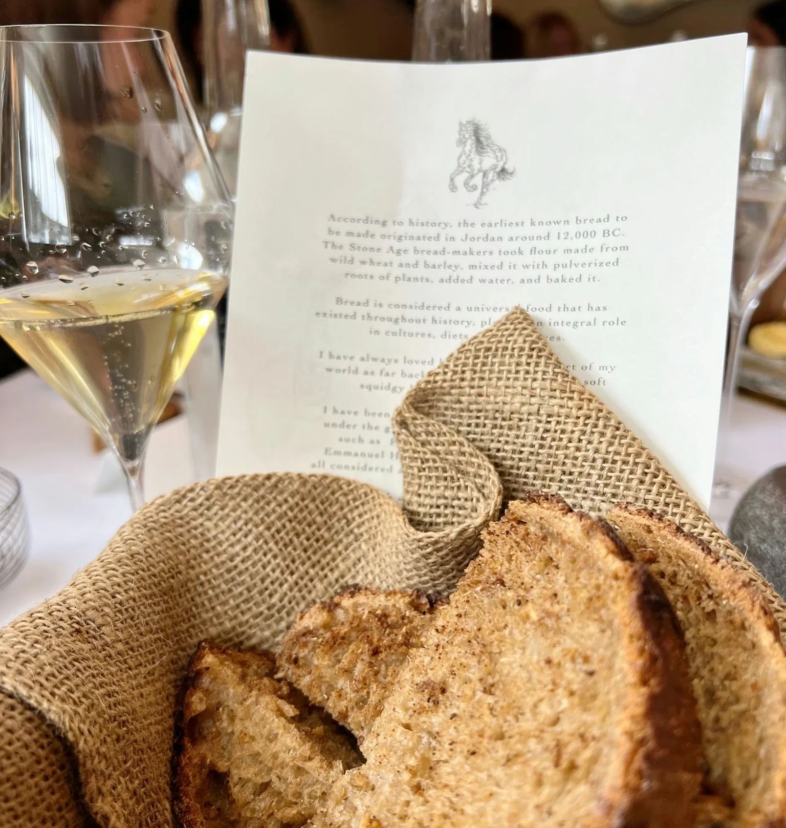 Sliced bread in a rustic cloth basket beside sparkling wine and a printed menu