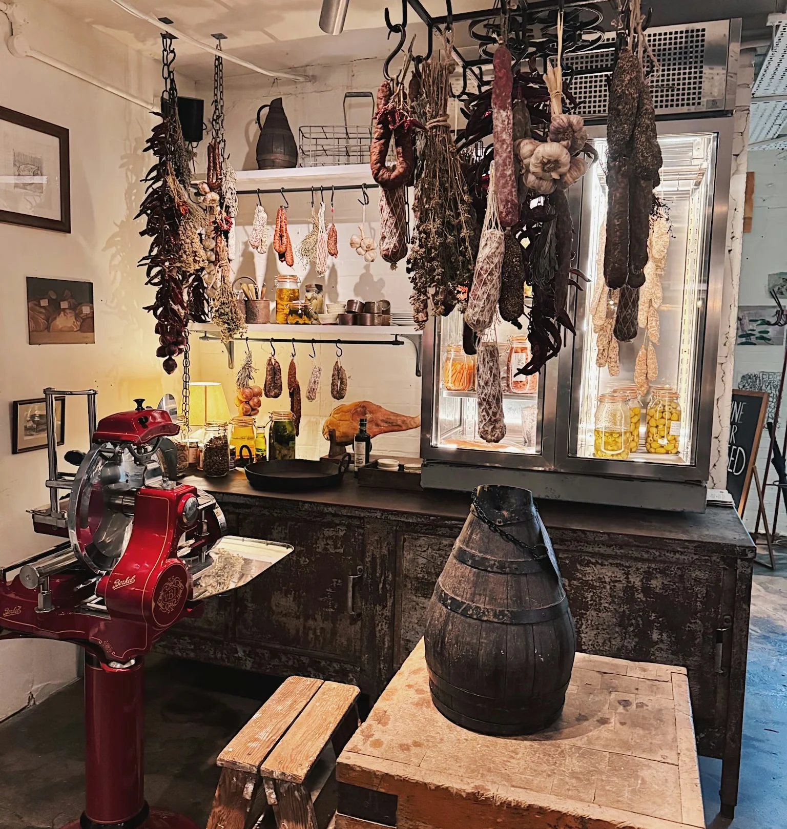 Traditional food shop interior with hanging cured meats, preserves, a red slicer and a wooden barrel