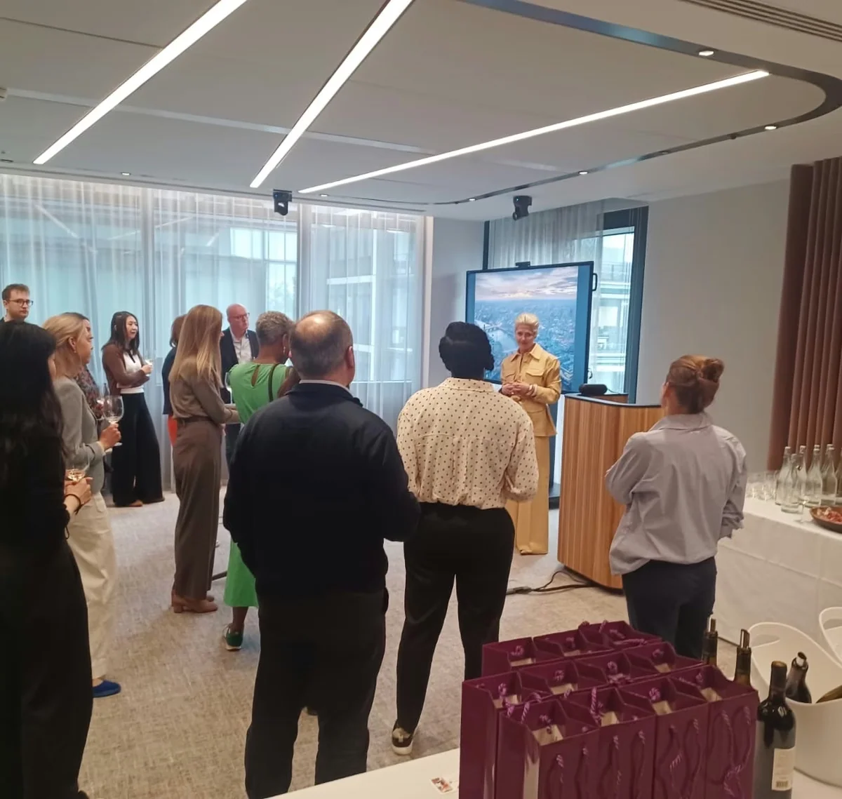 Guests gather around a speaker at a wine networking event, with wine glasses, bottles and refreshments on tables.