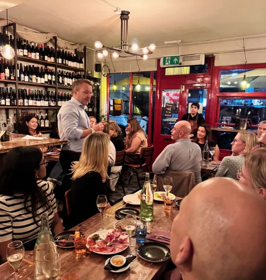 Wine tasting group gathered around tables as a man speaks in a busy restaurant
