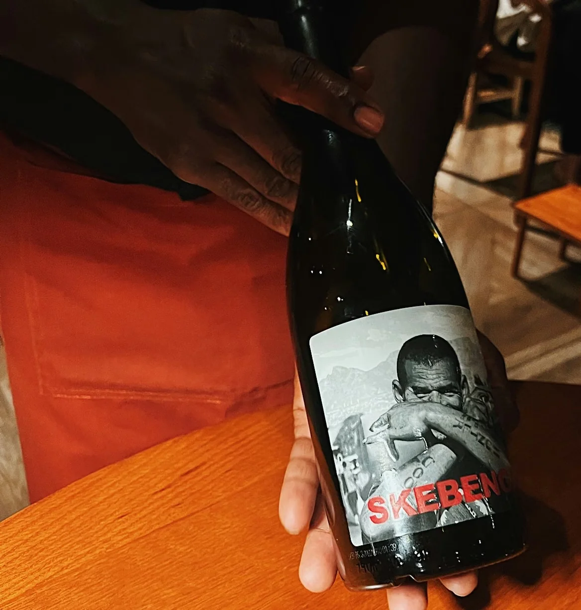 Hands holding a dark wine bottle with a black and white portrait label over a wooden table