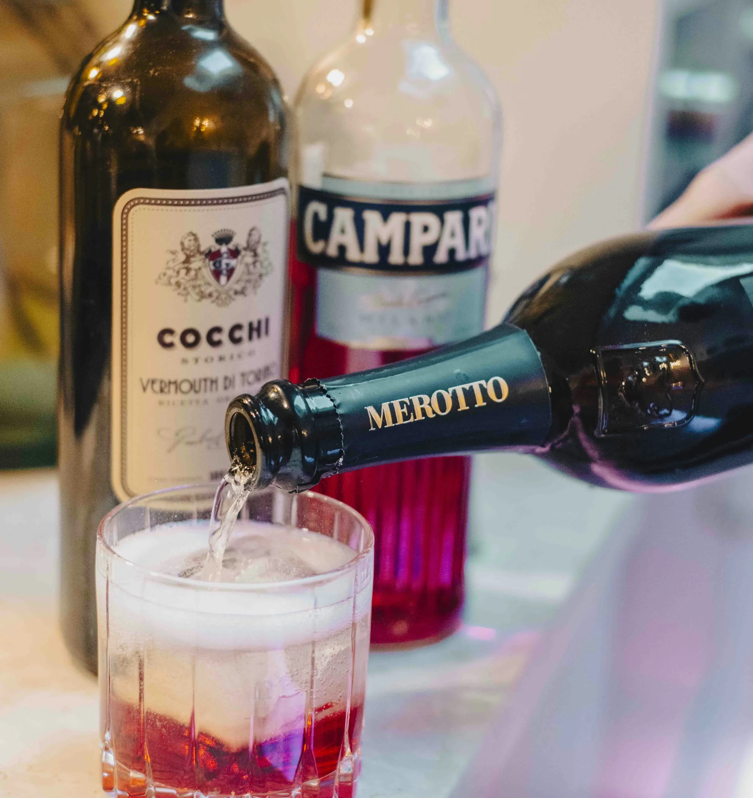 Prosecco poured into a red cocktail with Campari and Cocchi vermouth bottles behind