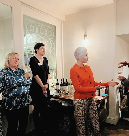 Guests listen to a speaker during an indoor wine tasting, with bottles and glasses arranged behind them