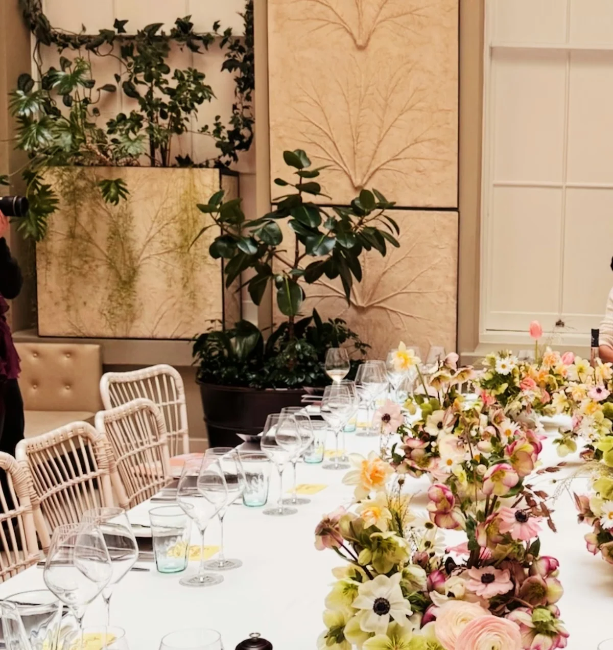 Long dining table set with wine glasses, pale rosé, floral centrepieces and leafy plants