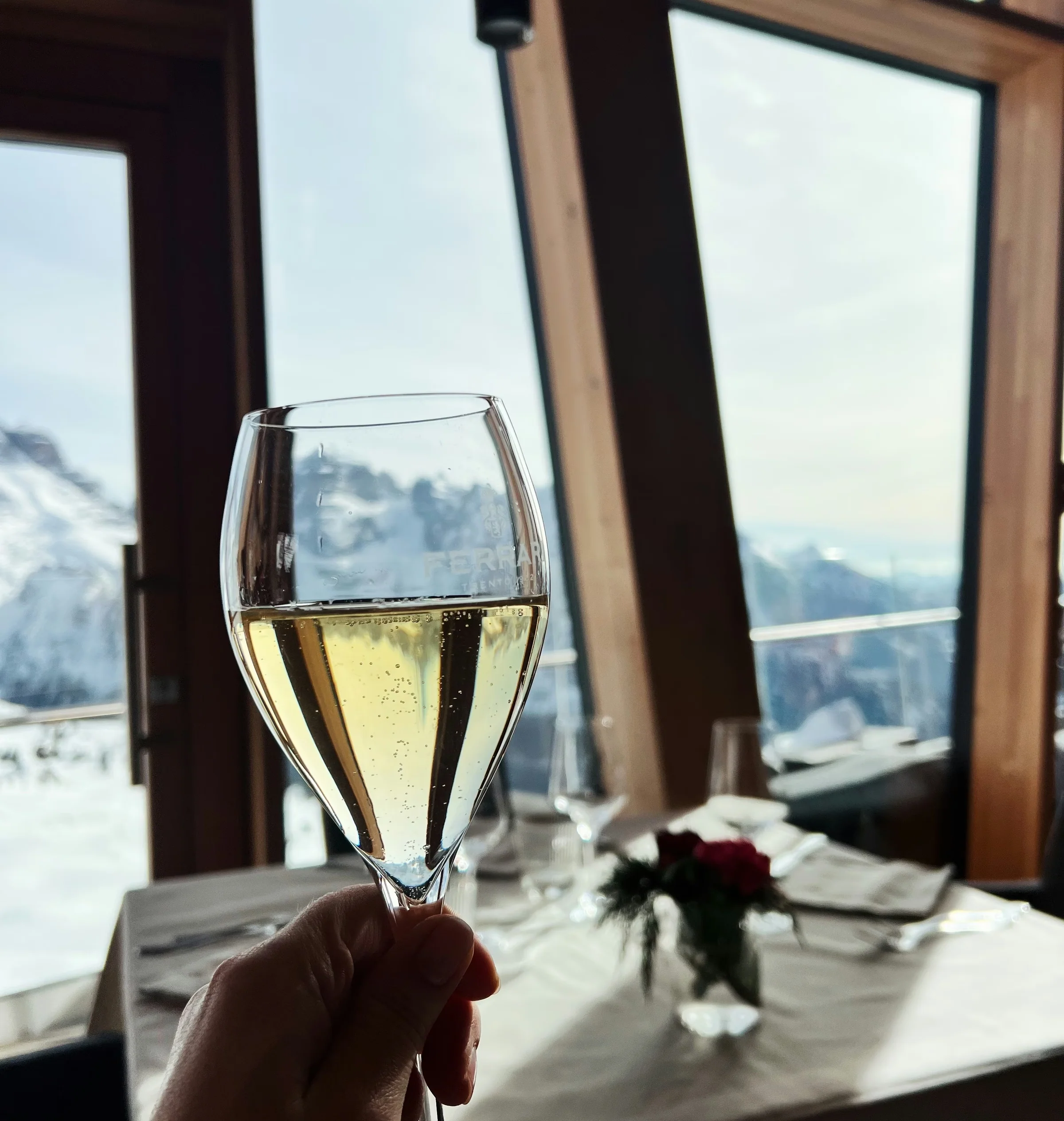 Hand holding a glass of sparkling wine beside a restaurant table, with snowy Dolomite mountains visible through the windows