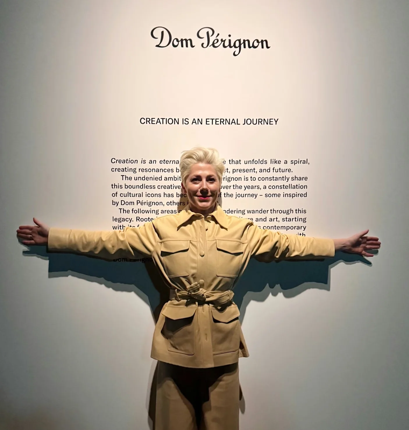 A woman in a belted beige outfit stands with arms out before a Dom Pérignon display at Tate Modern.