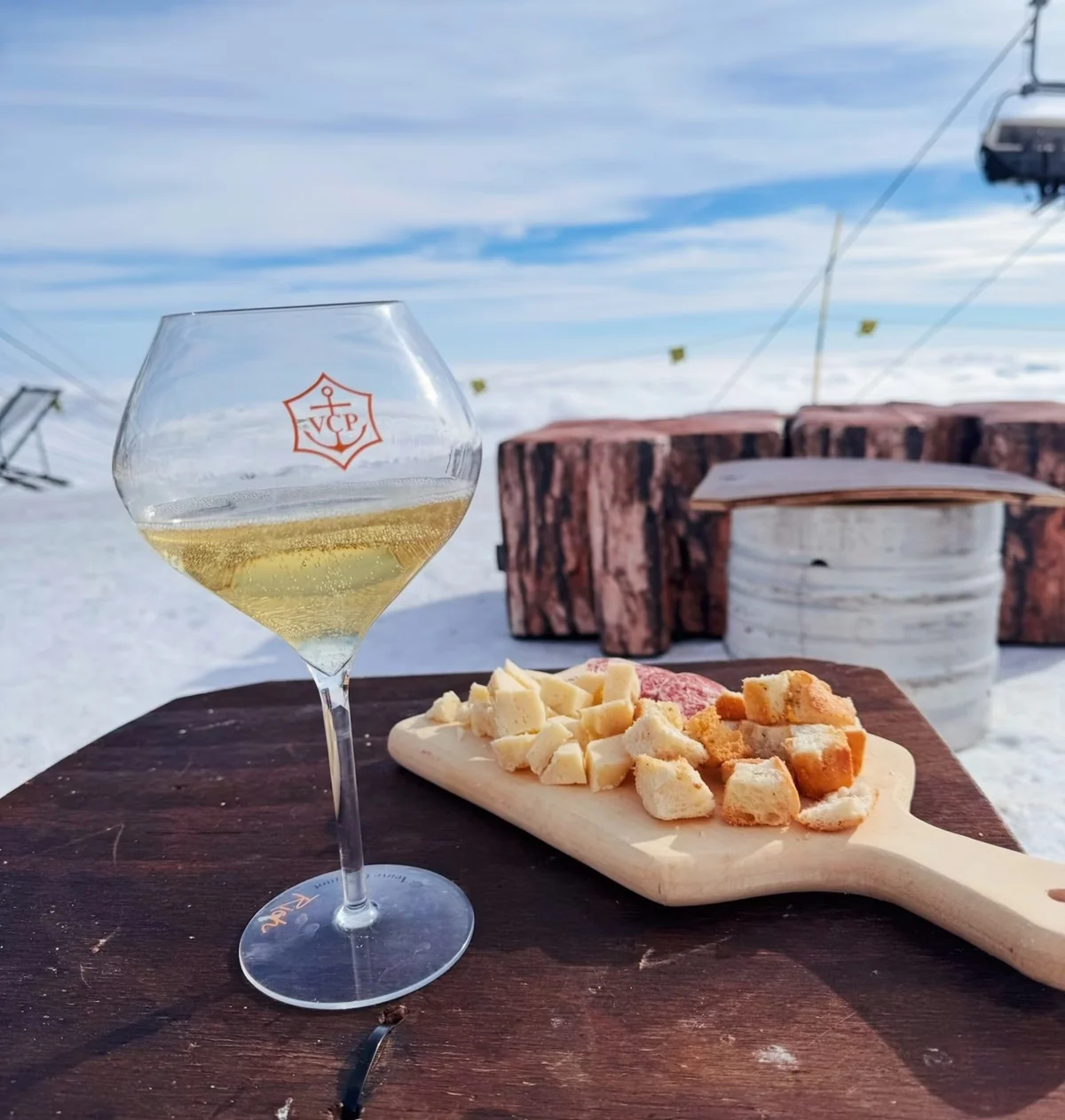 Glass of sparkling wine and a cheese and bread board on a mountainside terrace, with a ski lift behind