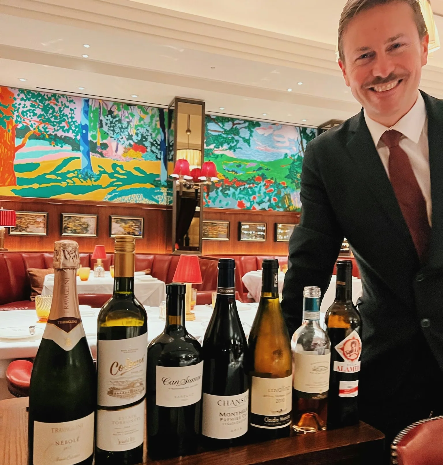 Man in a suit standing behind a display of wine bottles in a colourful restaurant