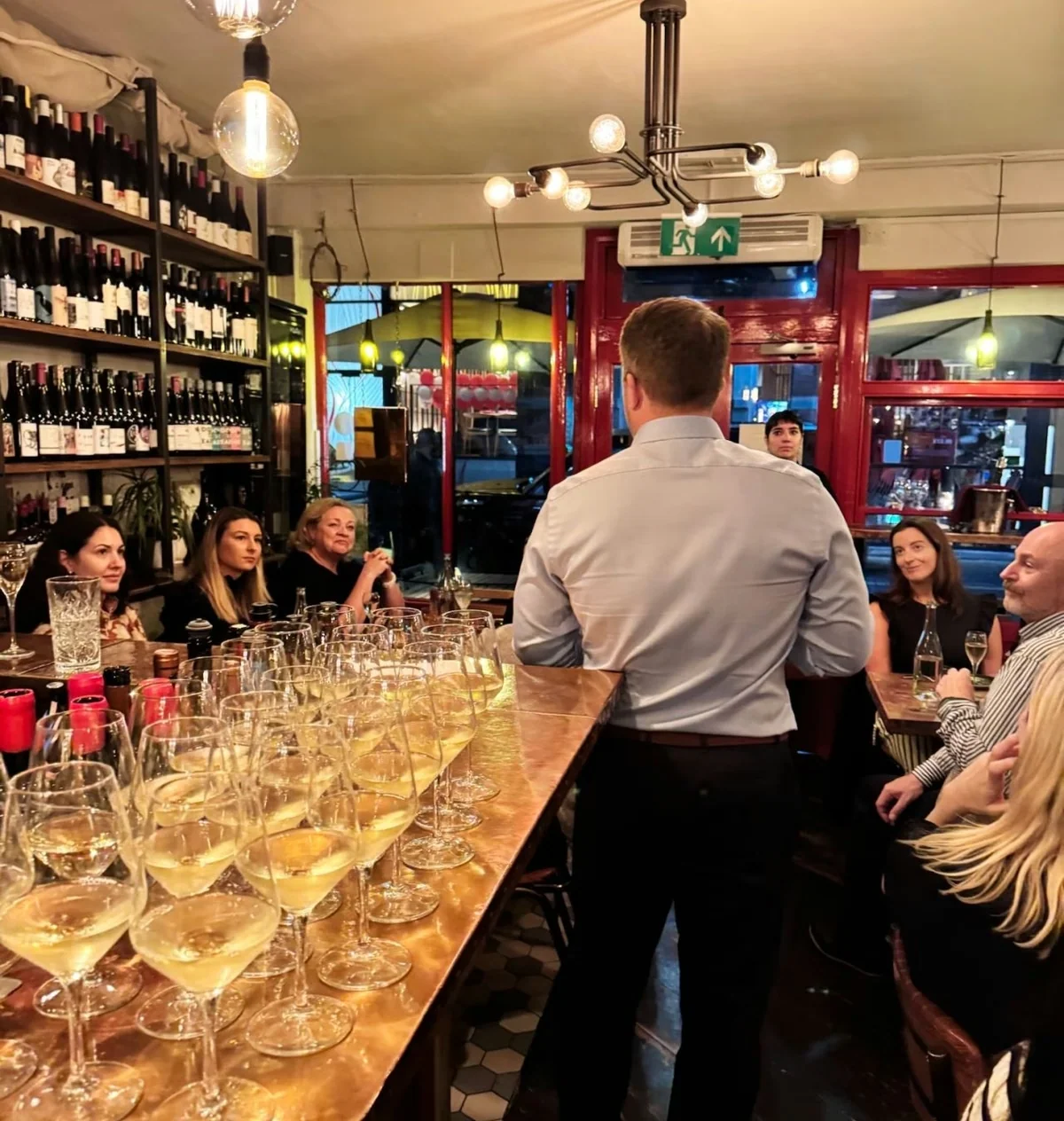 Wine tasting in a restaurant with a host speaking to guests around a bar lined with white wine glasses