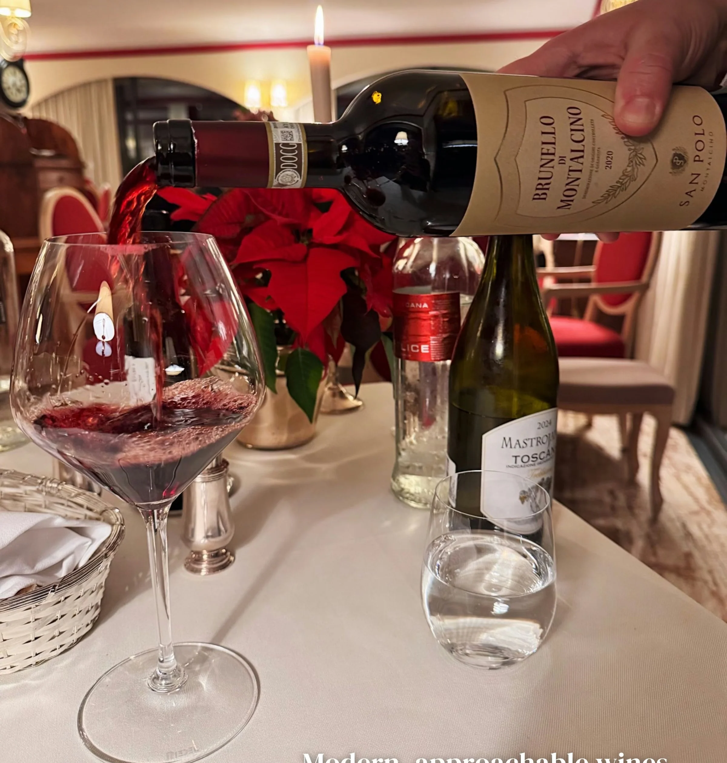 Red wine poured into a glass beside labelled Italian wine bottles on a candlelit restaurant table