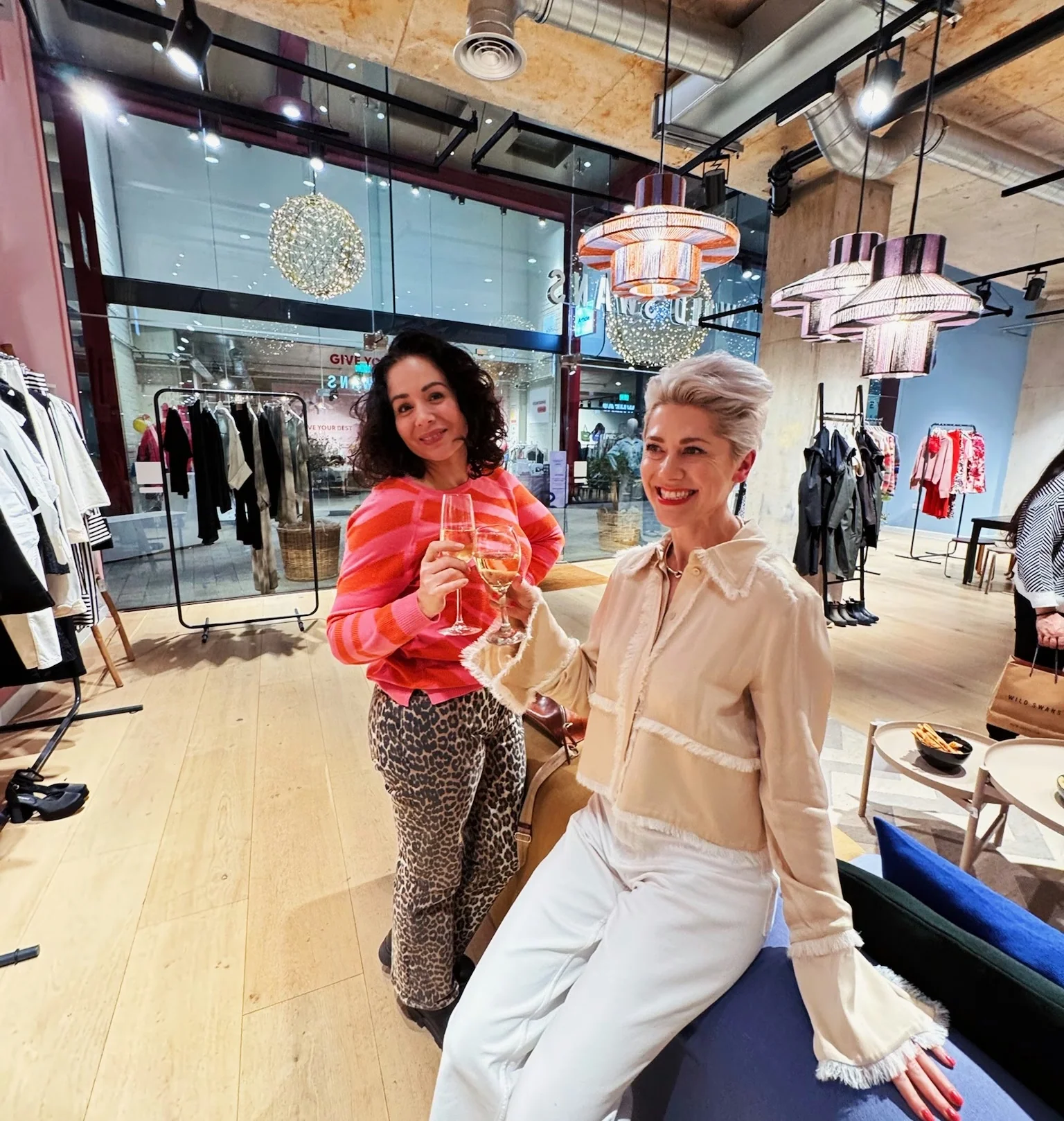 Two women holding glasses of pale wine in a stylish fashion showroom