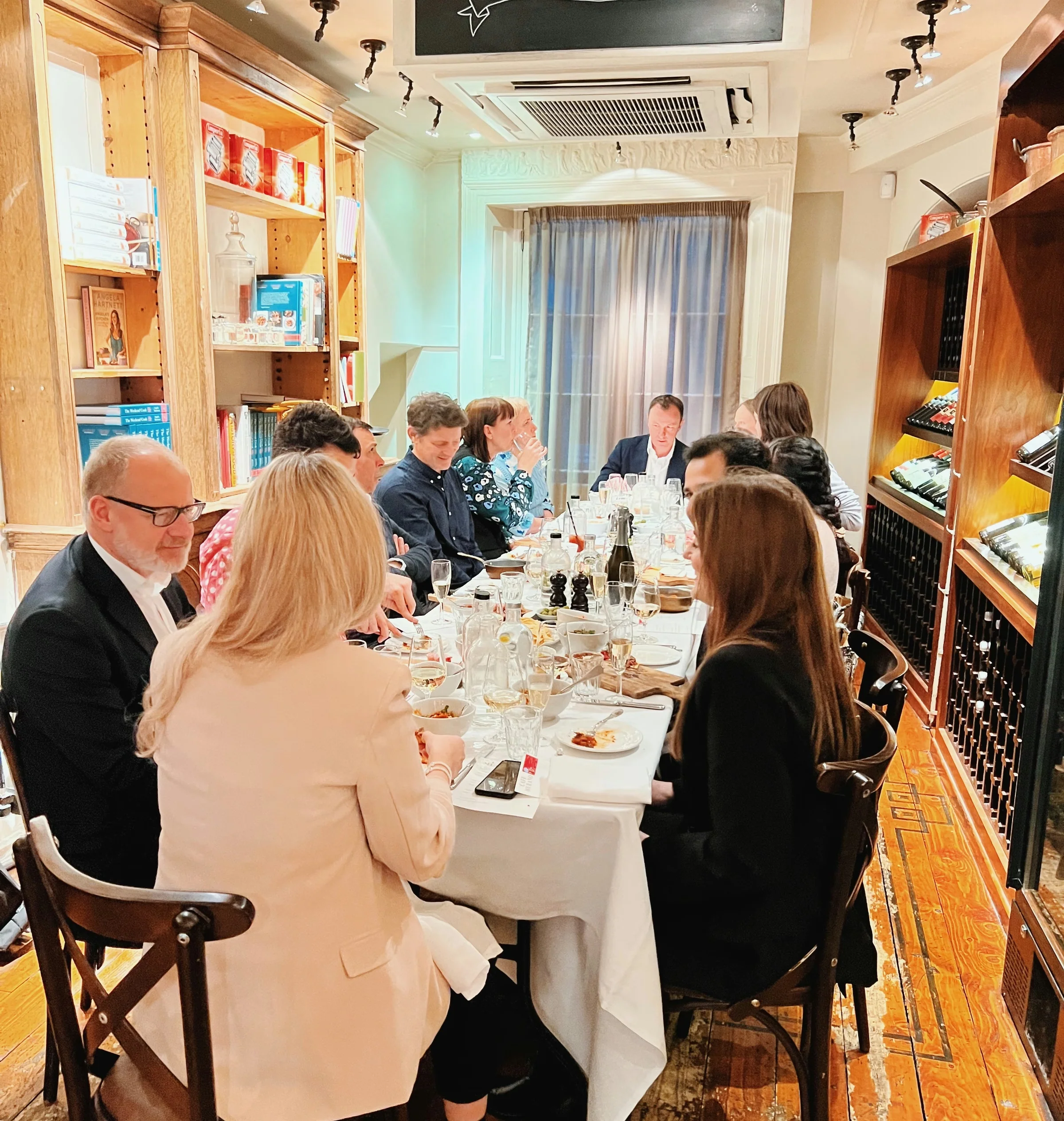 Guests share a pasta meal with wine at a long table in a restaurant