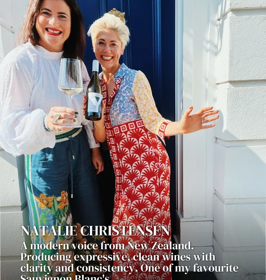 Two women at a doorway, holding a wine glass and bottle beneath text about Natalie Christensen