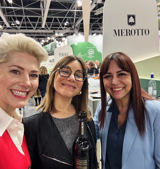 Three women pose with a bottle of Merotto wine at a busy wine exhibition stand.