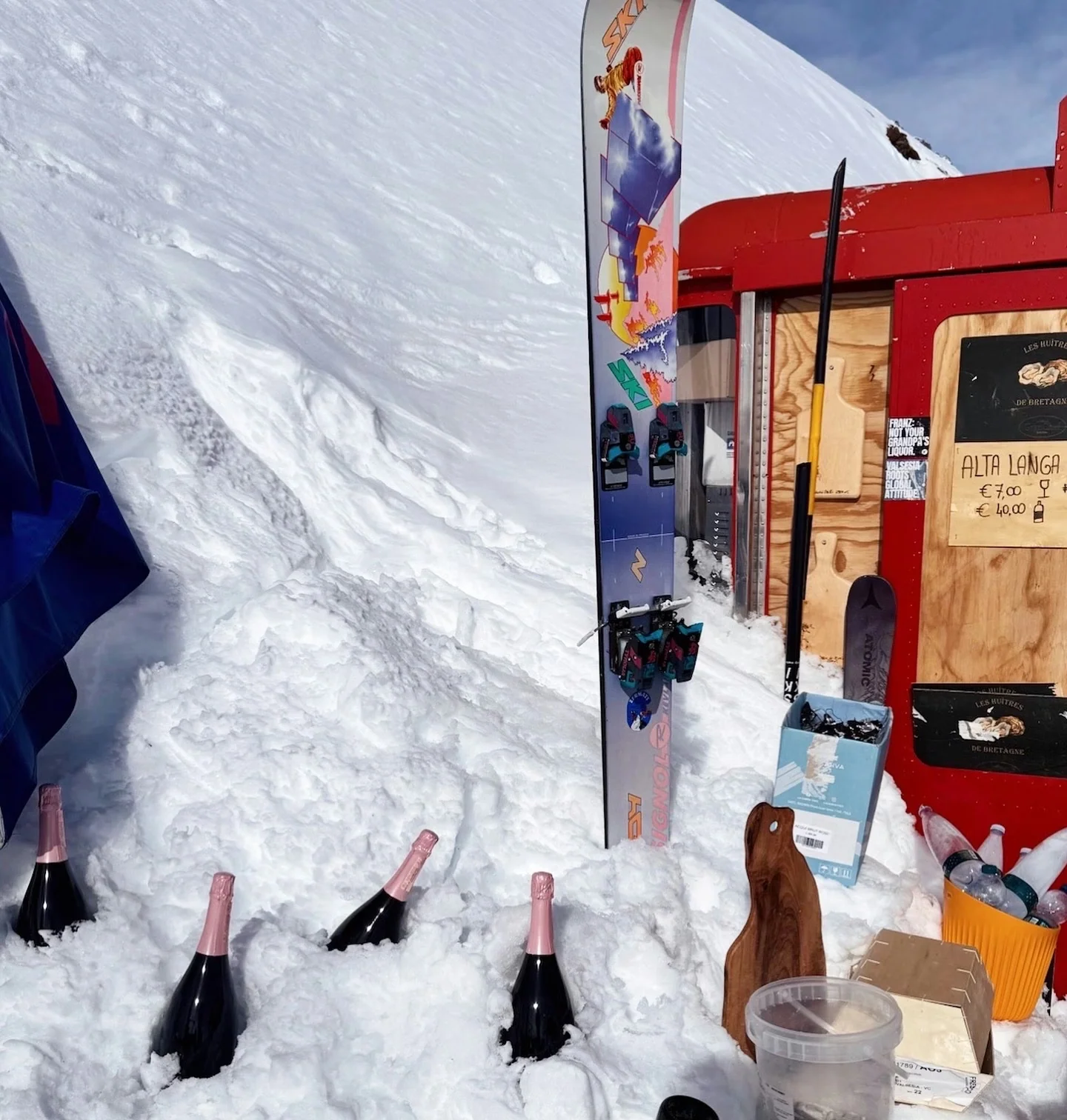 Champagne bottles chilled in snow outside a ski hut, with a snowboard and an Alta Langhe sign